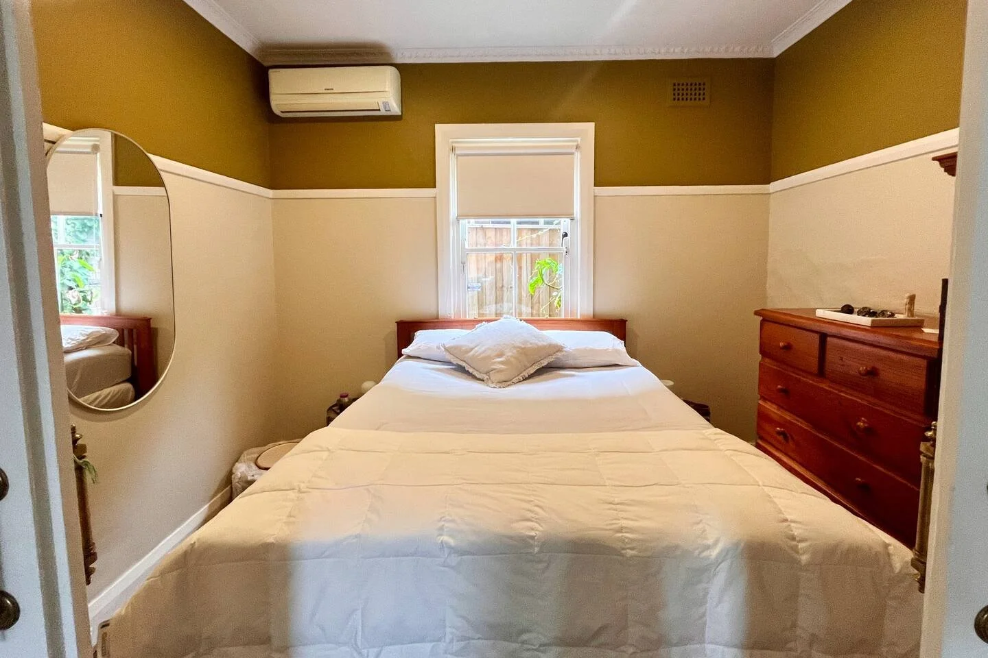 A bedroom with a bed, a window, a dresser, and an air conditioning unit, with a mirror on the wall.