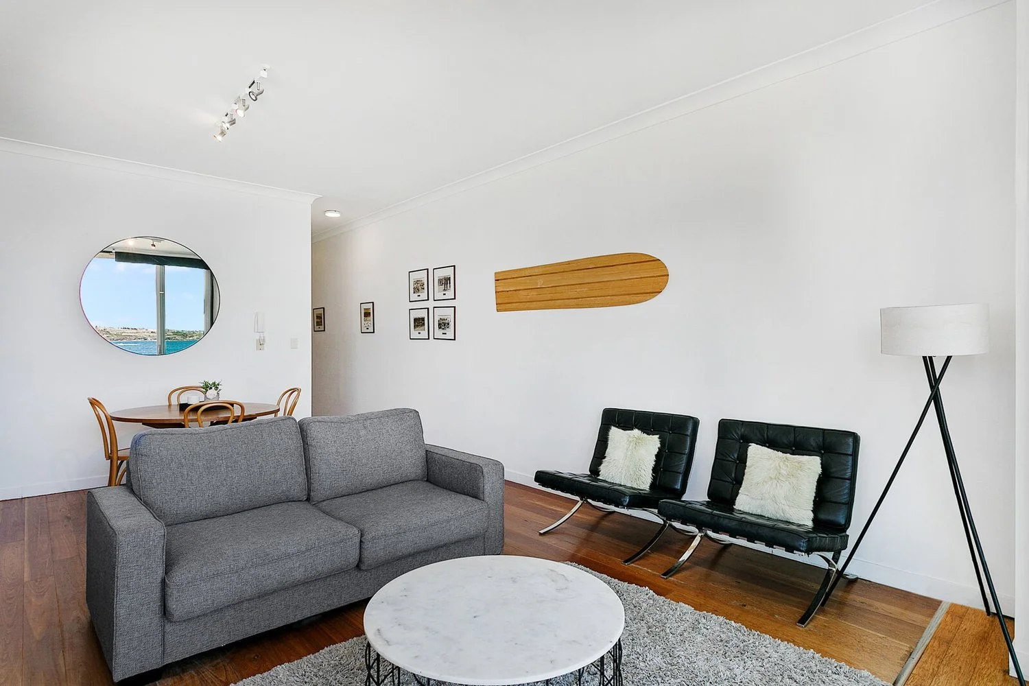 Modern living room with a gray couch, black leather chairs with white pillows, a round marble coffee table, and a tall tripod floor lamp. In the background, there's a small round dining table with four wooden chairs and a mirror on the wall reflectin