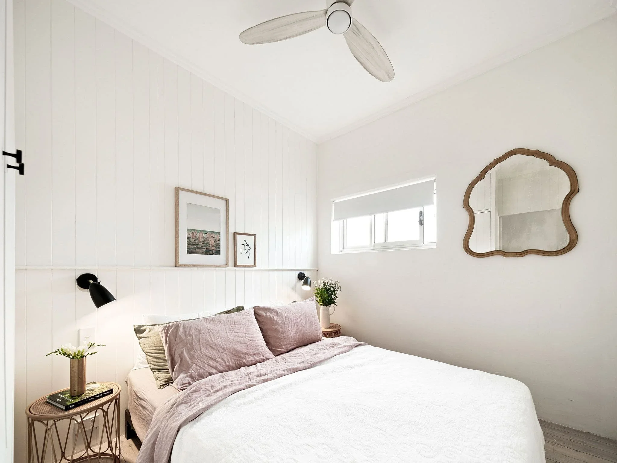 Minimalist bedroom with white walls, a small window, a wooden-framed mirror, two small pictures, a bed with pink and beige pillows, a black wall-mounted reading lamp, a plant on a nightstand, and a ceiling fan.