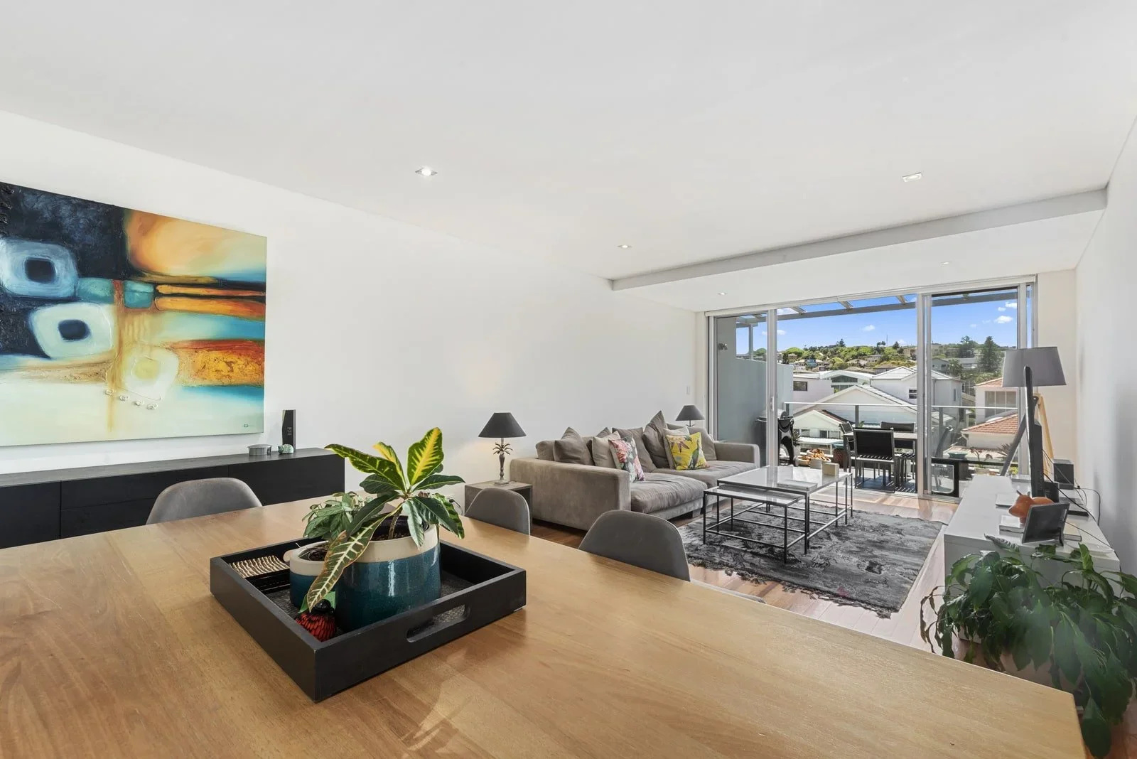 Modern living room with beige sofa, glass coffee tables, black and white area rug, and sliding glass doors leading to a balcony with outdoor furniture. Artwork on the white wall, potted plants on the dining table and near the TV, and a view of roofto