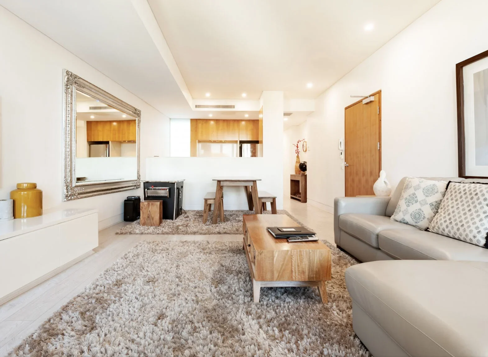 Bright living room with cream-colored walls, a large gray sectional sofa with patterned pillows, a wooden coffee table with magazines, a beige shag rug, a white media console, and a silver-framed mirror on the wall.