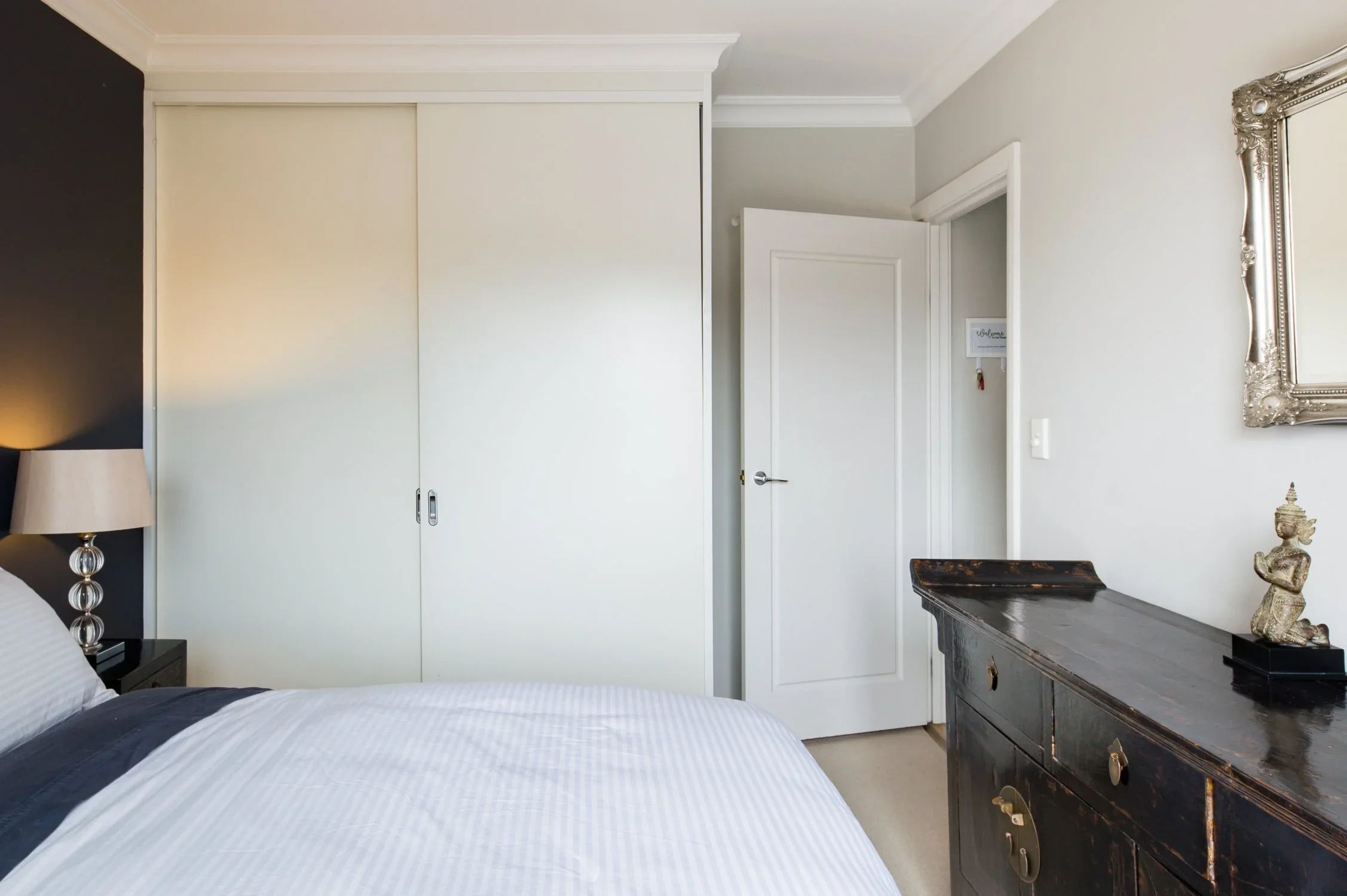 A bedroom with a bed, a nightstand with a lamp, a large white closet, a door, a black antique dresser with decorative items, and a framed mirror on the wall.