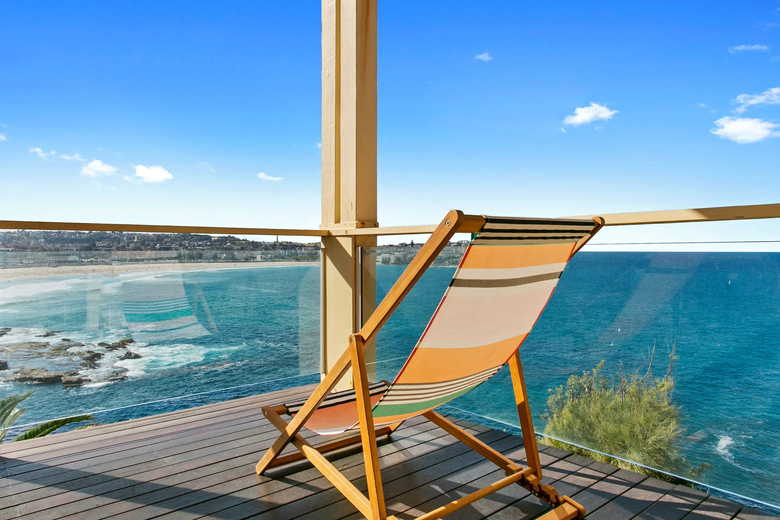 A deck chair on a wooden balcony overlooking the ocean with a sandy beach and cityscape in the background.