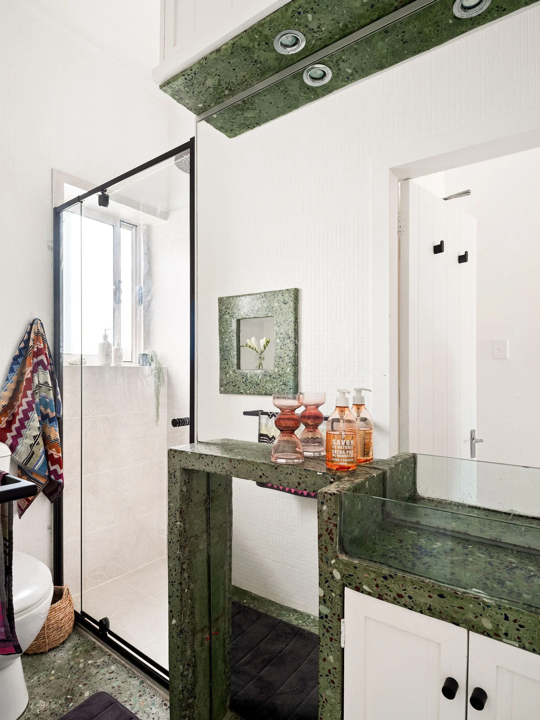 Modern bathroom with green terrazzo sink, mirror, glass bottles, walk-in shower with glass door, window, and minimal decor.