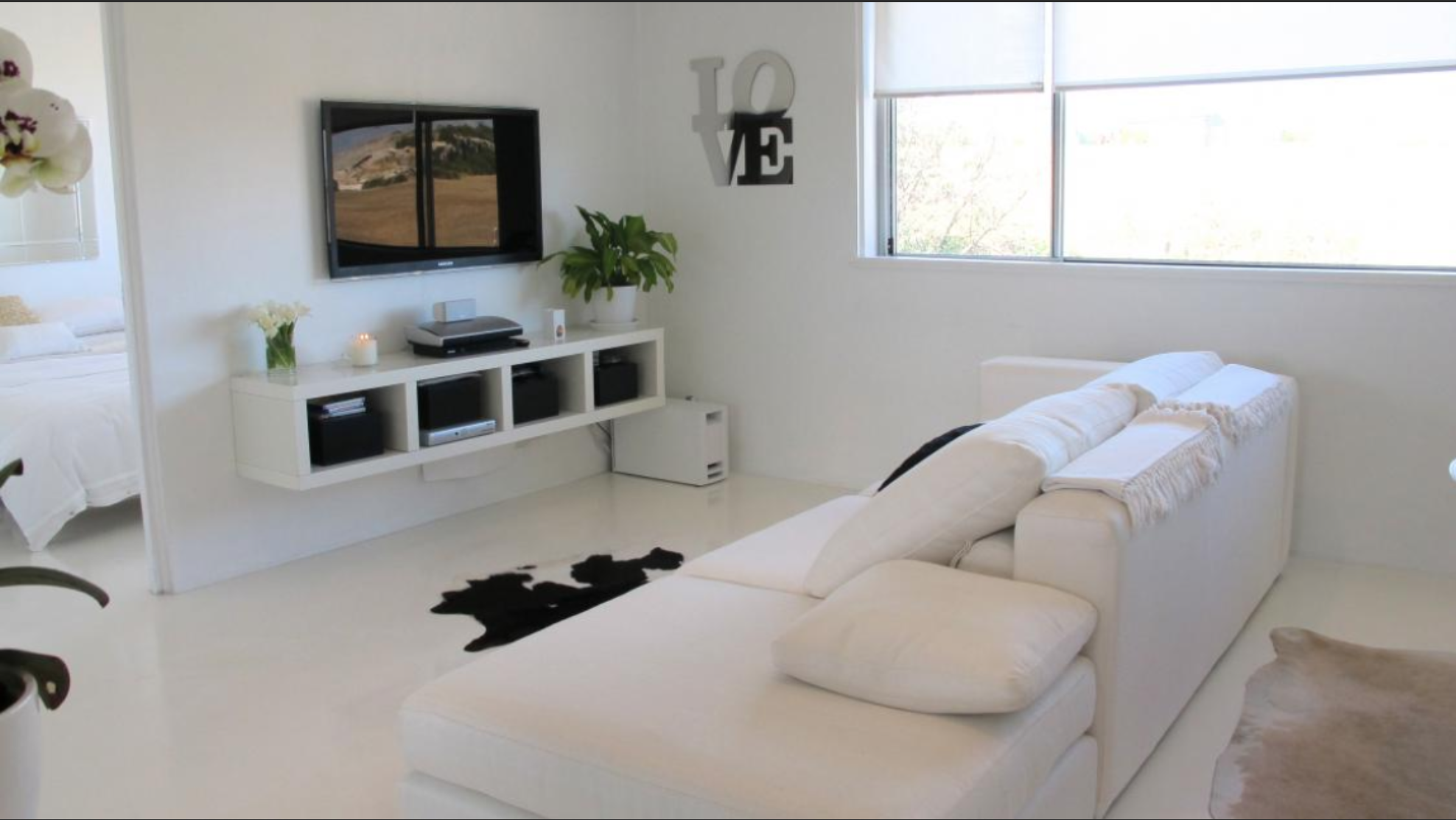 Bright living room with white sofa, wall-mounted TV, white entertainment unit, potted plant, decorative wall art spelling love, large window, and bedroom visible to the side.