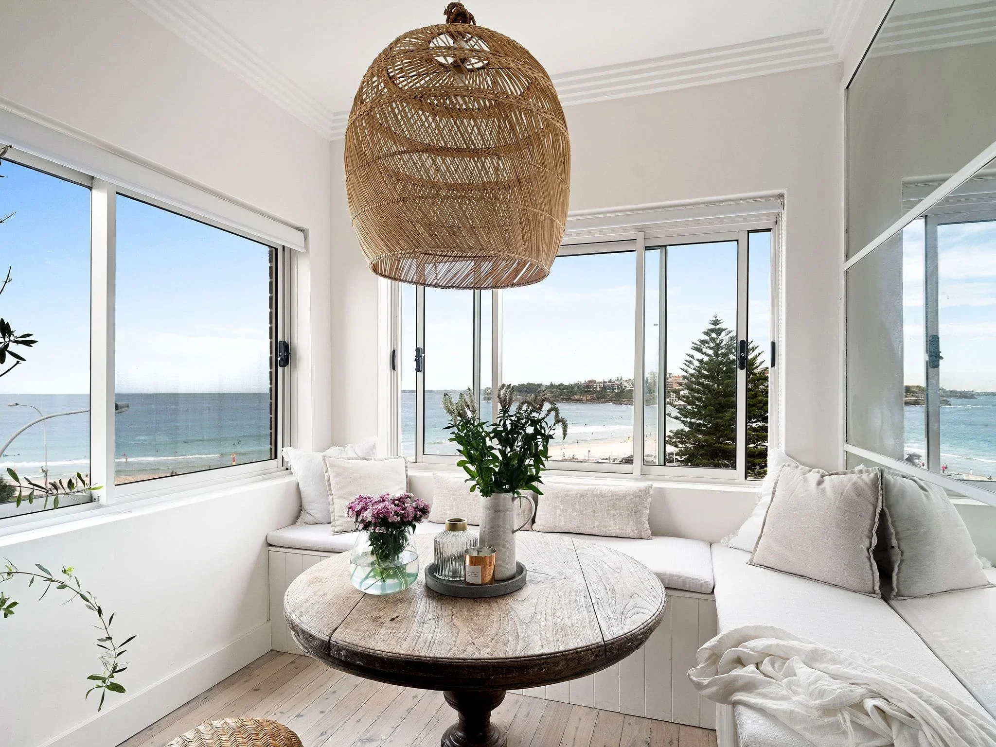 Cozy coastal sitting area with large windows overlooking the beach, white seating with cushions, a round wooden table with flowers, a large hanging wicker light, and a scenic ocean view.