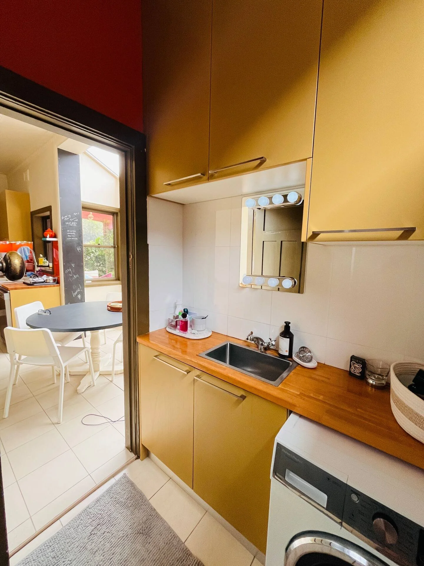 Small kitchen with yellow cabinets, a small sink, and a washing machine. A doorway leads to a dining area with a round table and white chairs, and a chalkboard wall is visible in the background.
