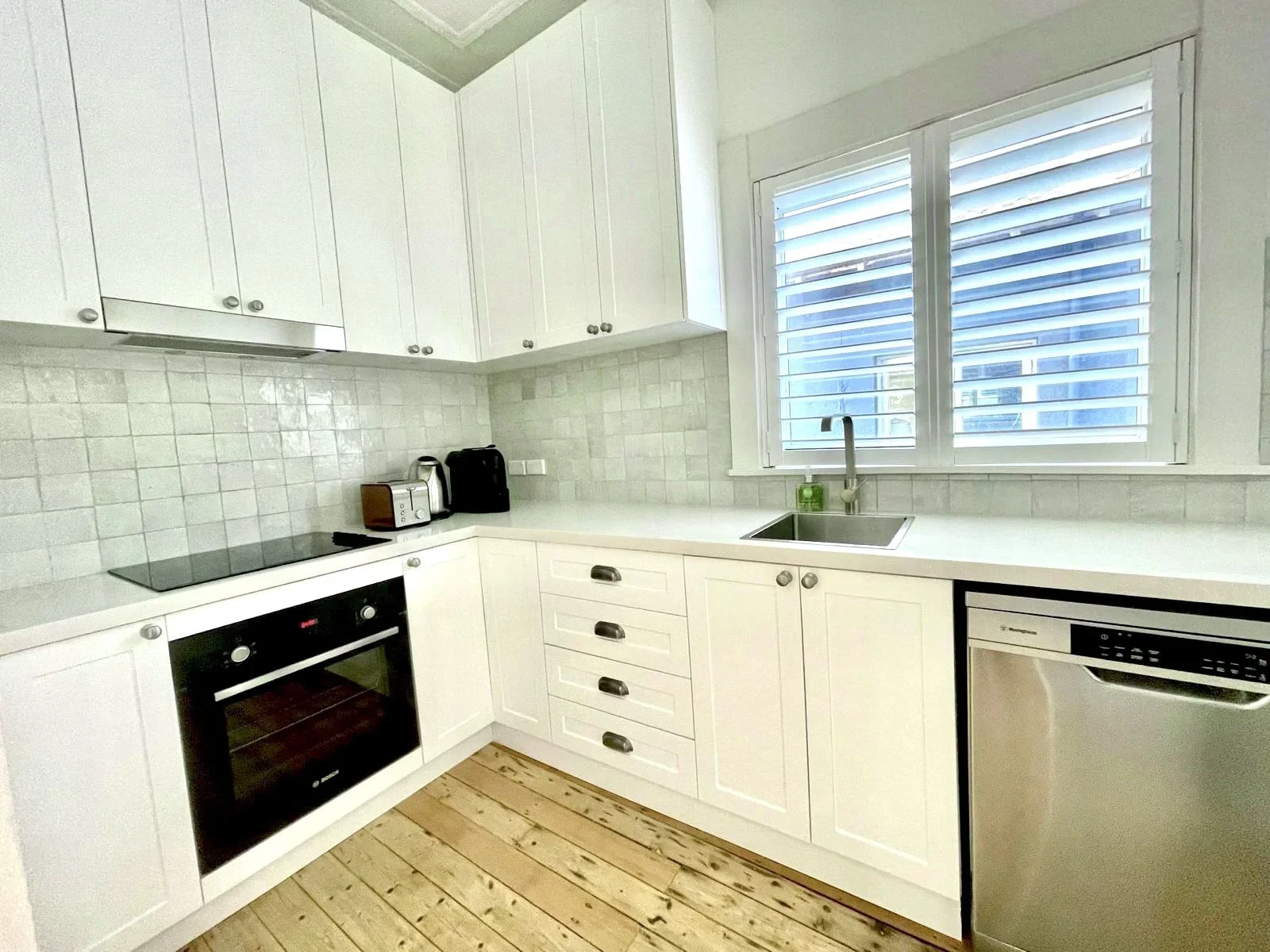 Bright white kitchen with white cabinets, a black oven, a washing machine, and a stainless steel sink under a window with white shutters.