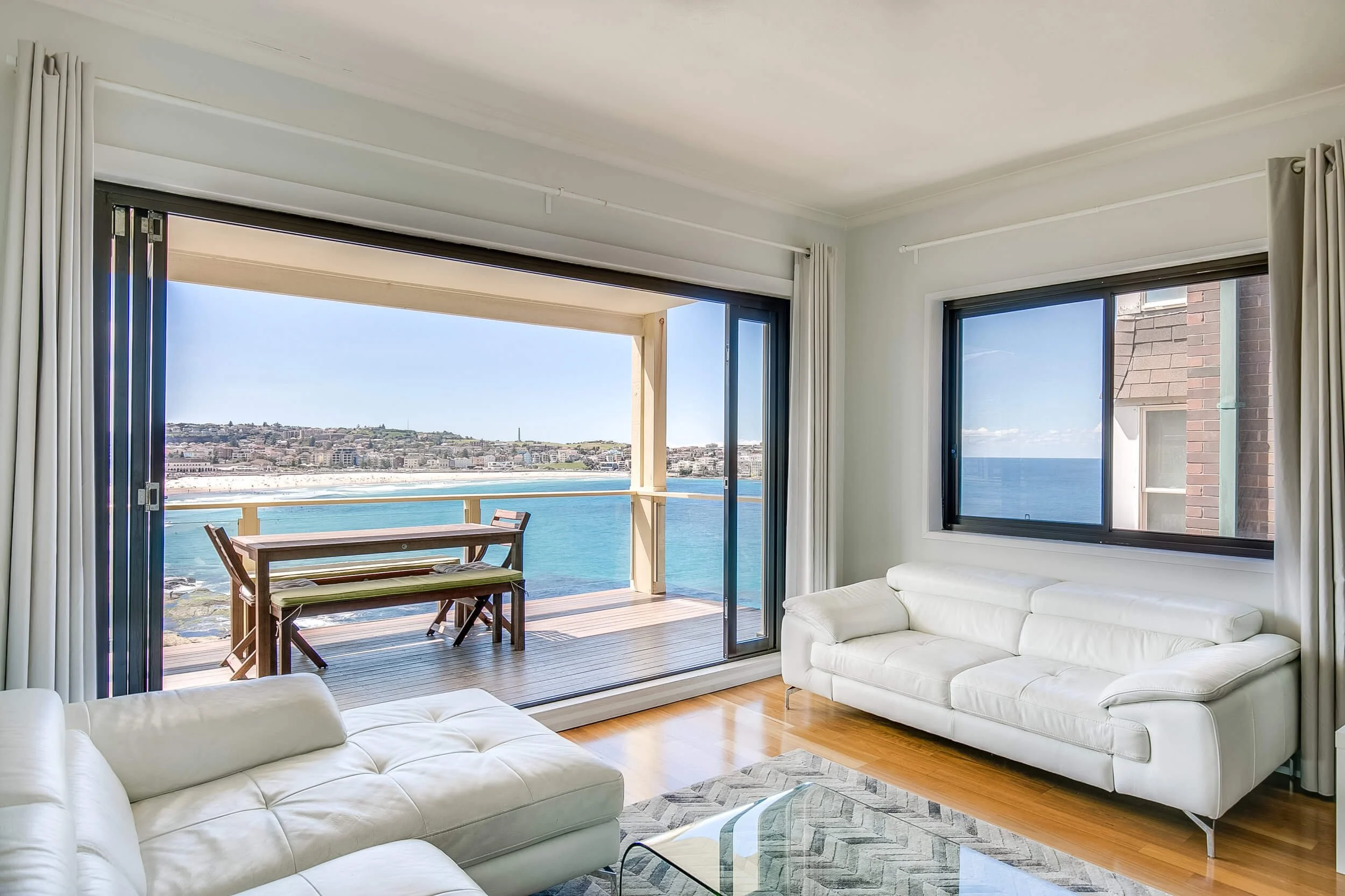 Living room with white sofas, large window showing ocean view, sliding glass door leading to a balcony with outdoor table and chairs, and wooden flooring.