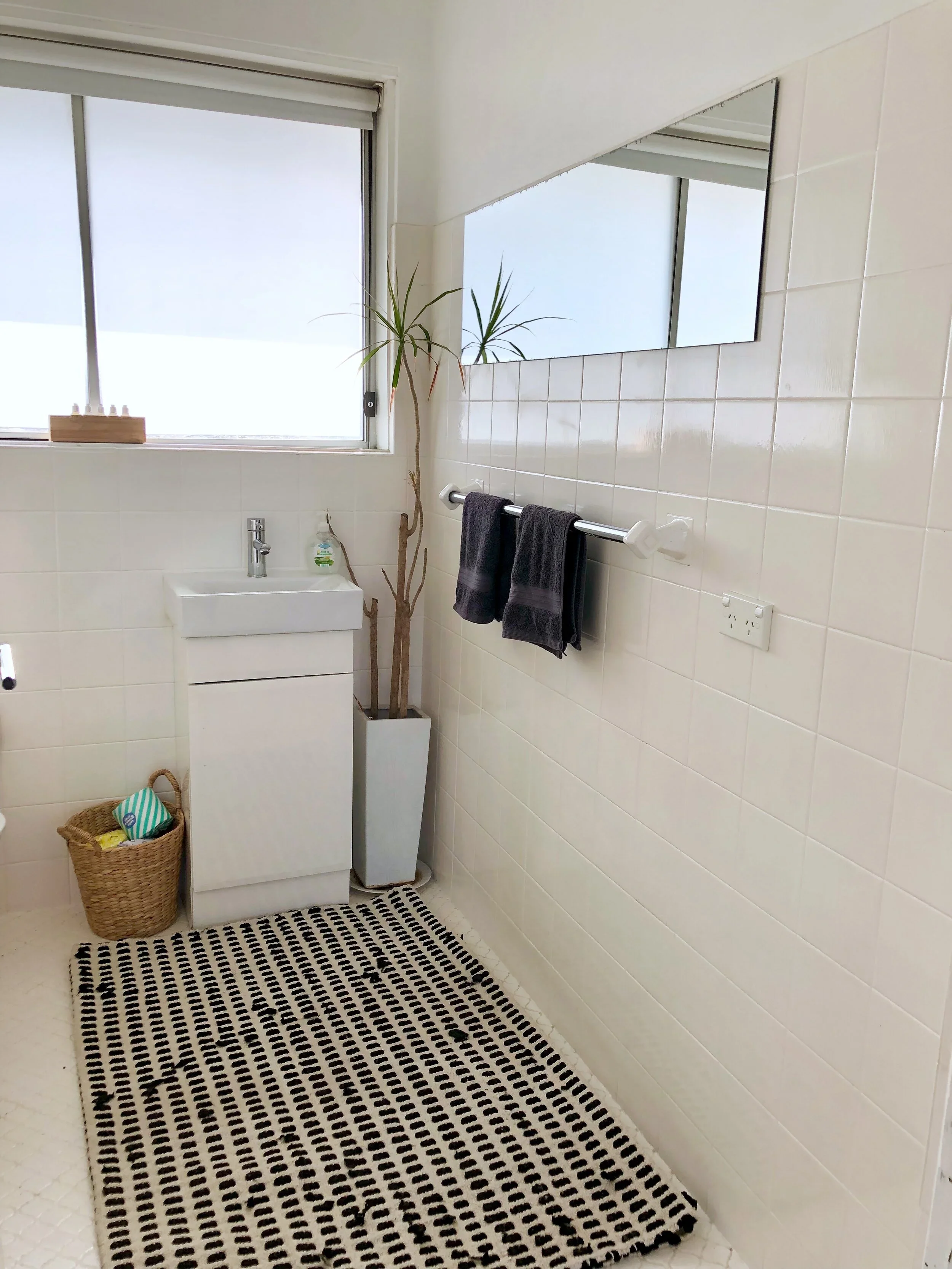 A minimalist bathroom with a white sink cabinet, a large mirror, two black towels on a silver towel rack, a potted plant, a window with a roller blind, a wicker basket, and a black and white striped rug on the floor.