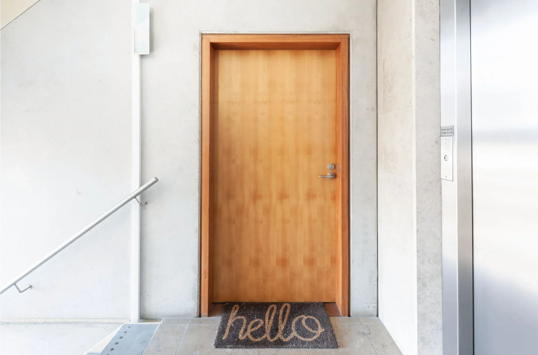 A closed wooden door in a building with a hello doormat in front of it, next to an elevator and a white wall with a handrail.