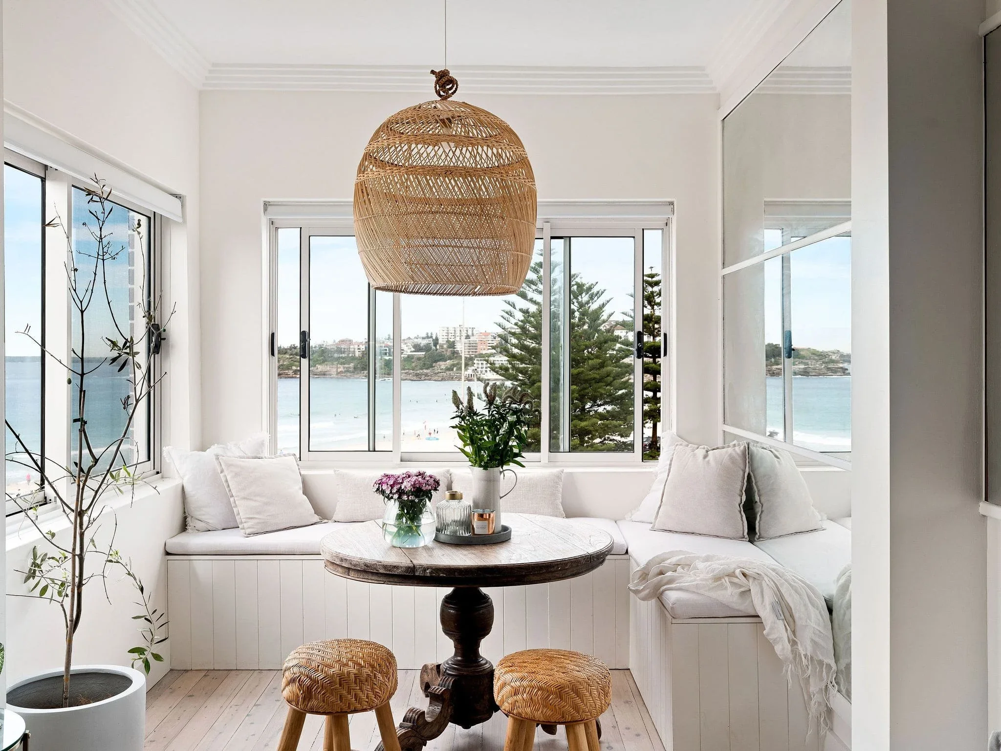Beach view through large windows in a bright, white sunroom with a round wooden table, two woven stools, plants, pillows, and a rattan pendant light.
