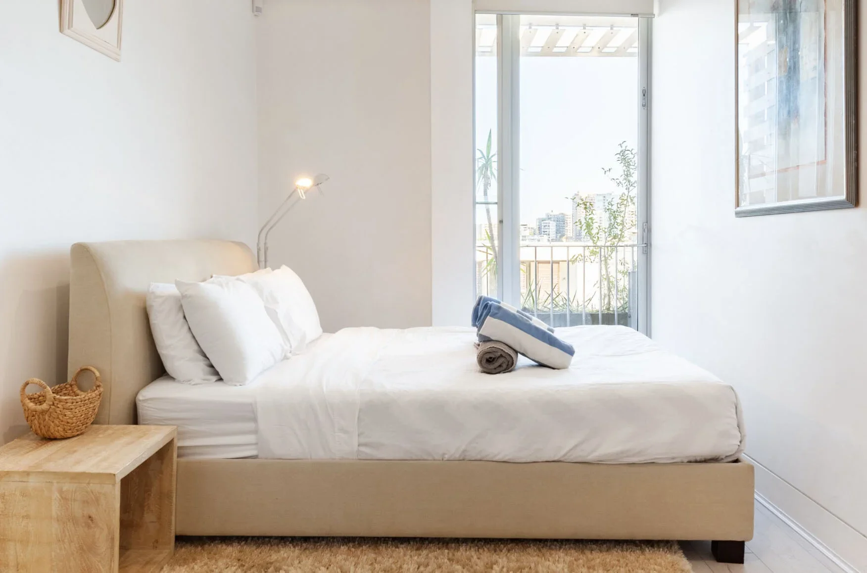 A neatly made bed with white bedding and pillows in a bright, modern bedroom. There are rolled towels on the bed, a small wooden nightstand with a wicker basket, and a sliding glass door leading to a balcony with city buildings and trees outside.