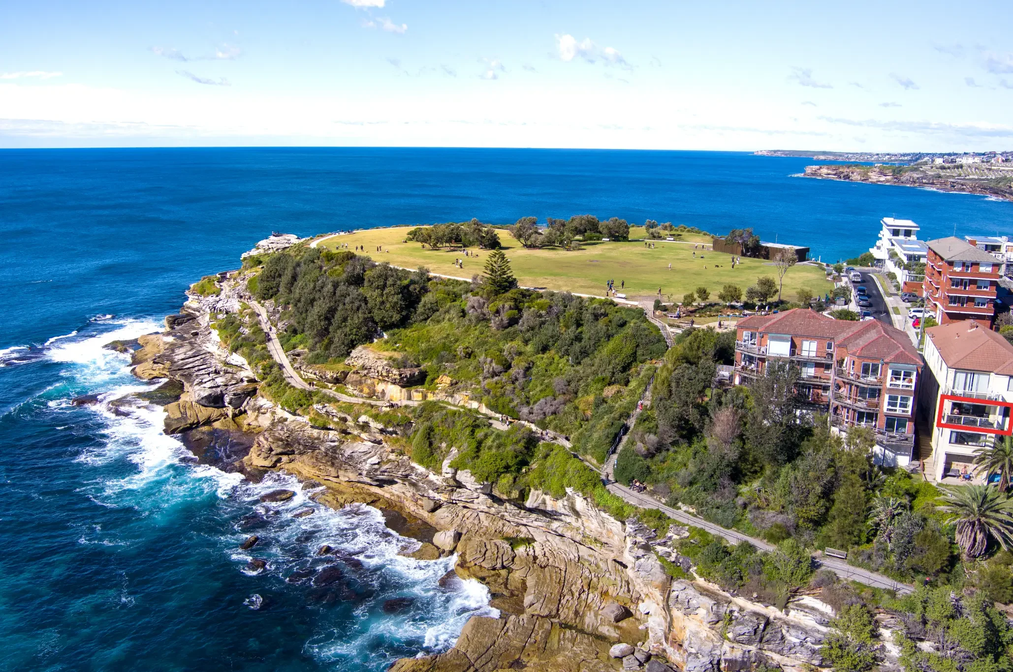 A coastal park with rocky cliffs, green grass, and walking paths overlooking the ocean, with buildings on the hillside.