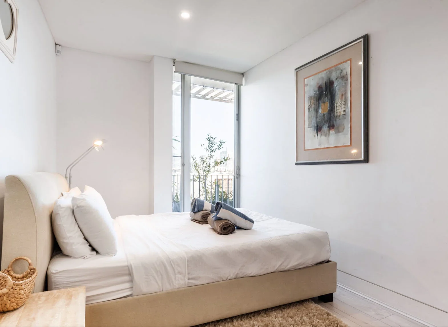 A bedroom with a beige upholstered bed, white bedding, and two rolled towels on the bed. There is a small wicker basket on a wooden nightstand. A modern abstract painting hangs on the white wall. Natural light enters through a sliding glass door that