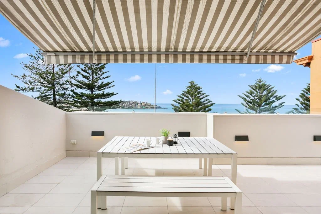 Balcony with white outdoor furniture, overlooking pine trees and ocean view under a striped awning.