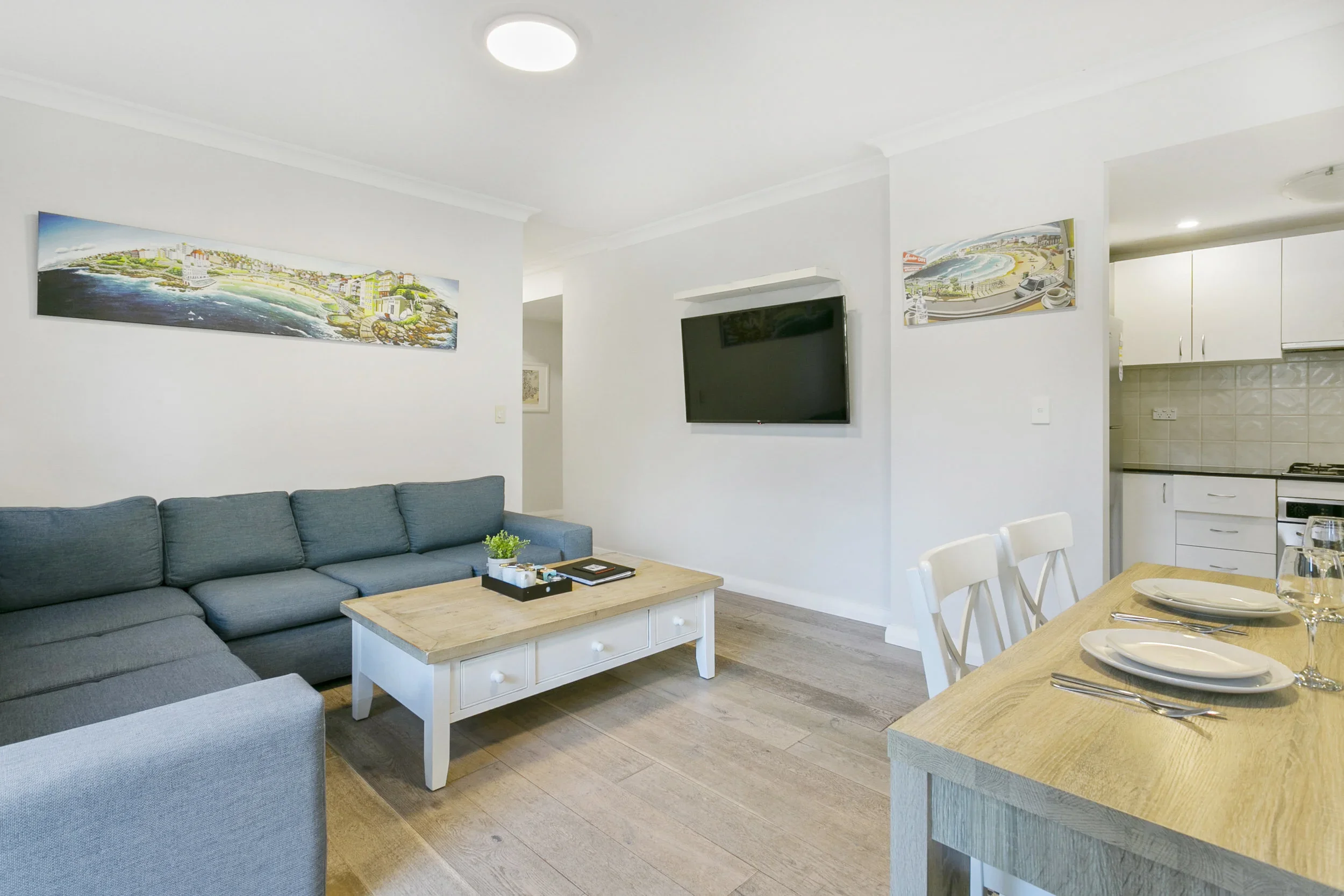 Living room with a large gray sectional sofa, a white coffee table, wall-mounted TV, and wall art of cityscapes. Adjacent to a dining area with a wooden table set with plates and glasses, and a view into a white kitchen with cabinets and appliances.
