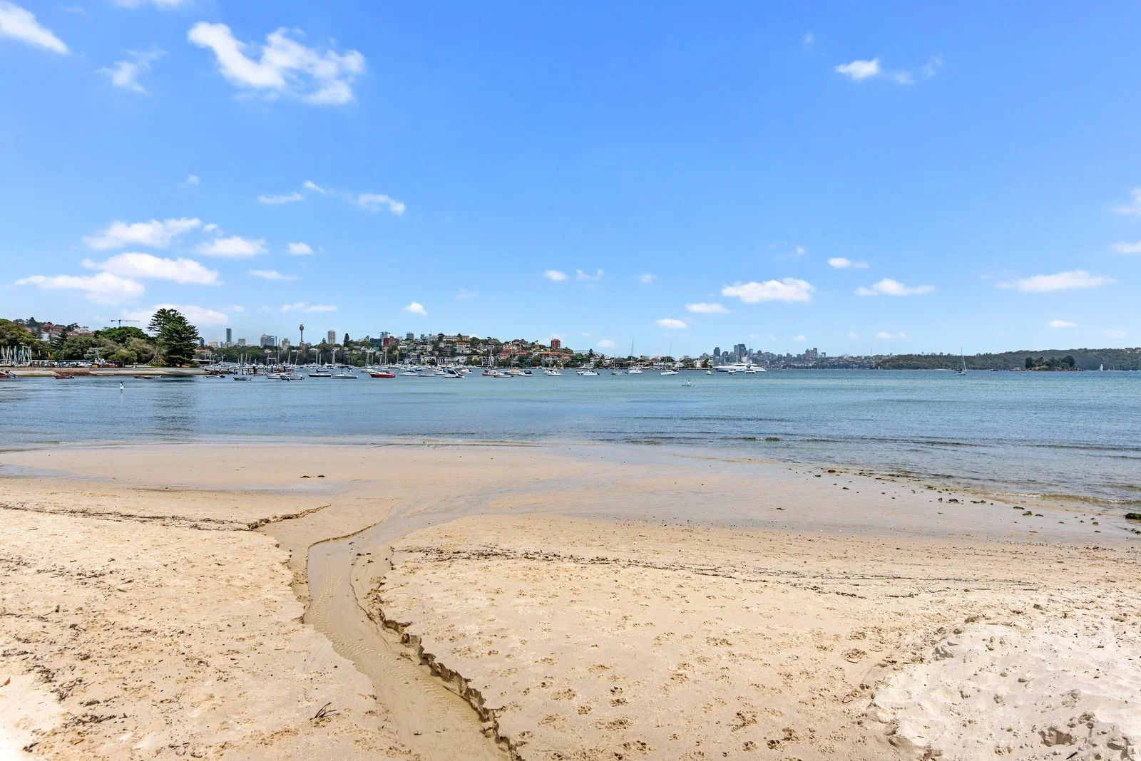 A sandy beach leading to calm water with numerous boats anchored near the shoreline. In the background, an urban skyline with various buildings and a clear blue sky with scattered clouds.
