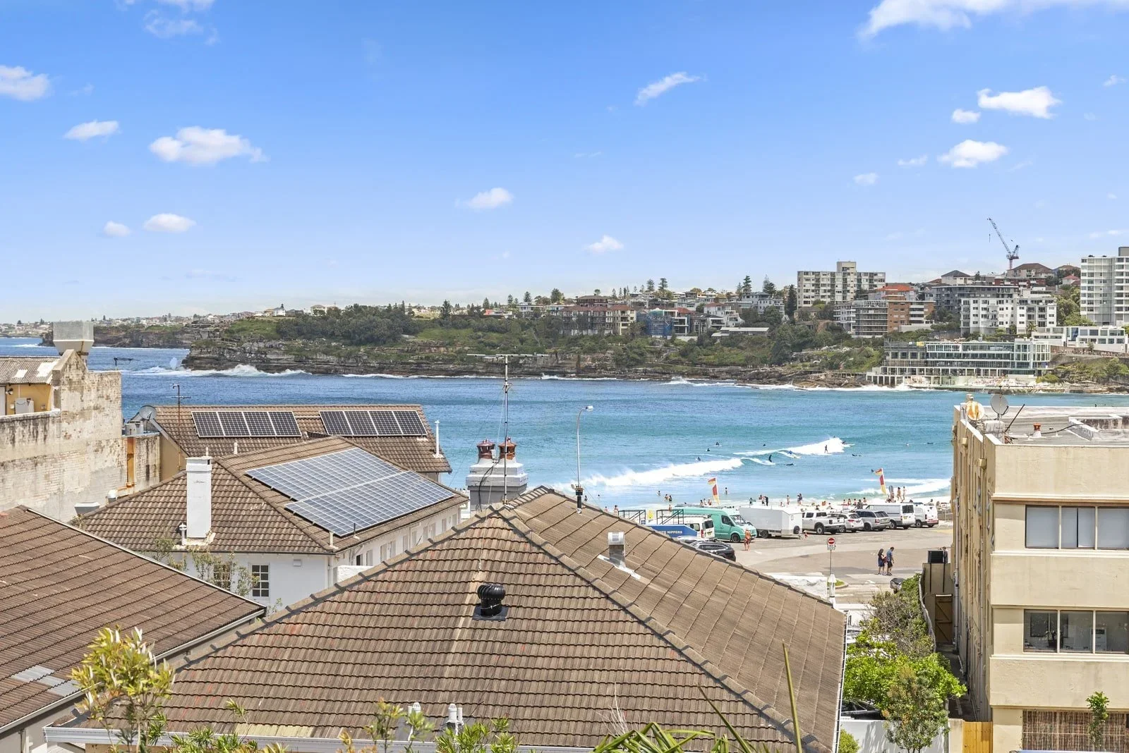 View of a coastal city with rooftops, a beach with people and boats, and a skyline of high-rise buildings under a blue sky with a few clouds.
