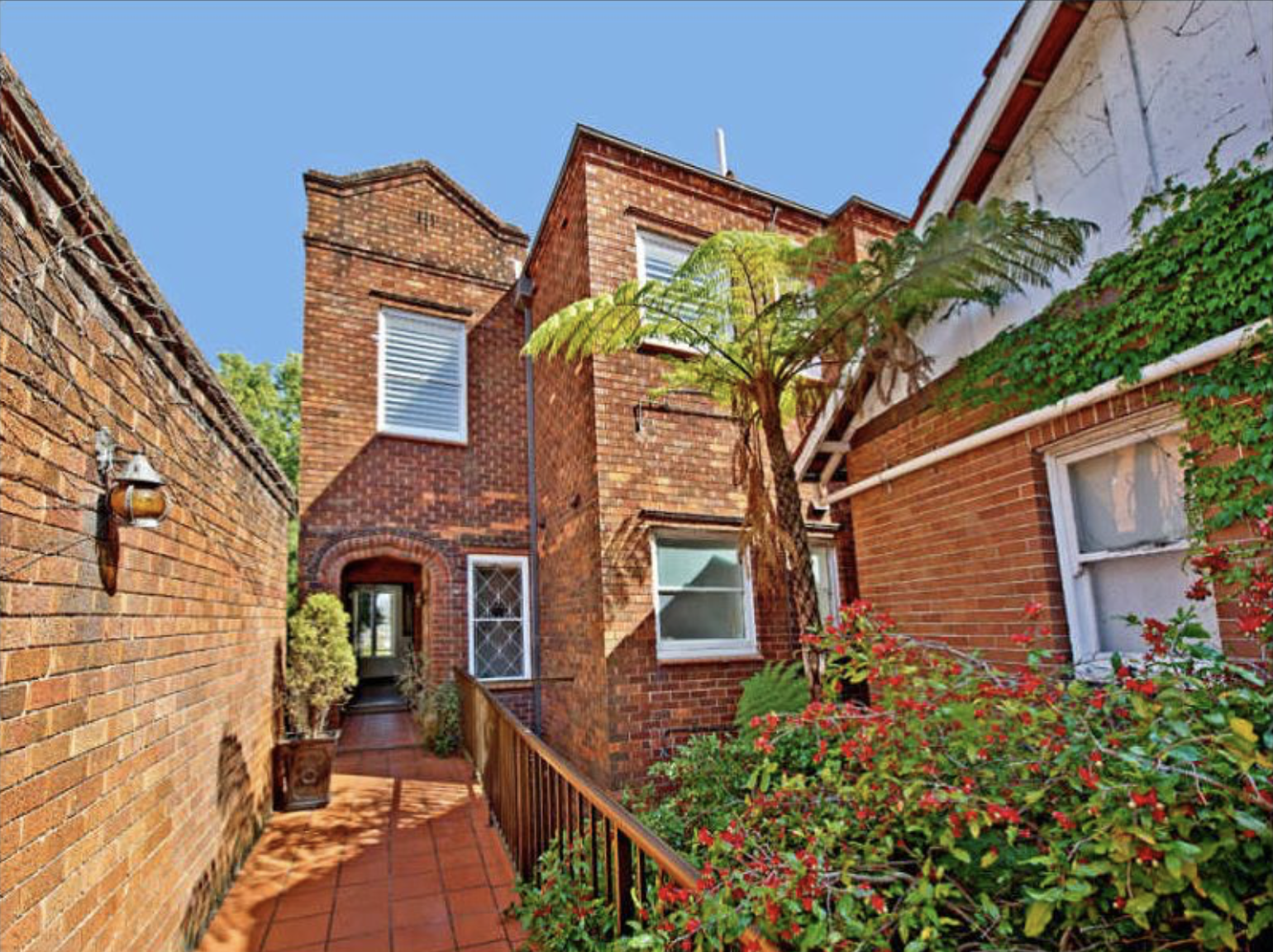 A brick alleyway with plants and an arched doorway leading into a brick building under a clear blue sky.