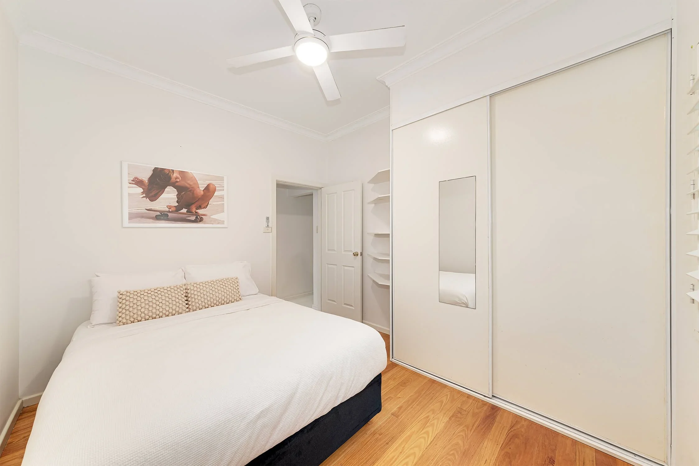 Minimalist bedroom with white bed, wall art of a skateboarder, built-in white closet with sliding doors, a mirror, and wooden flooring.