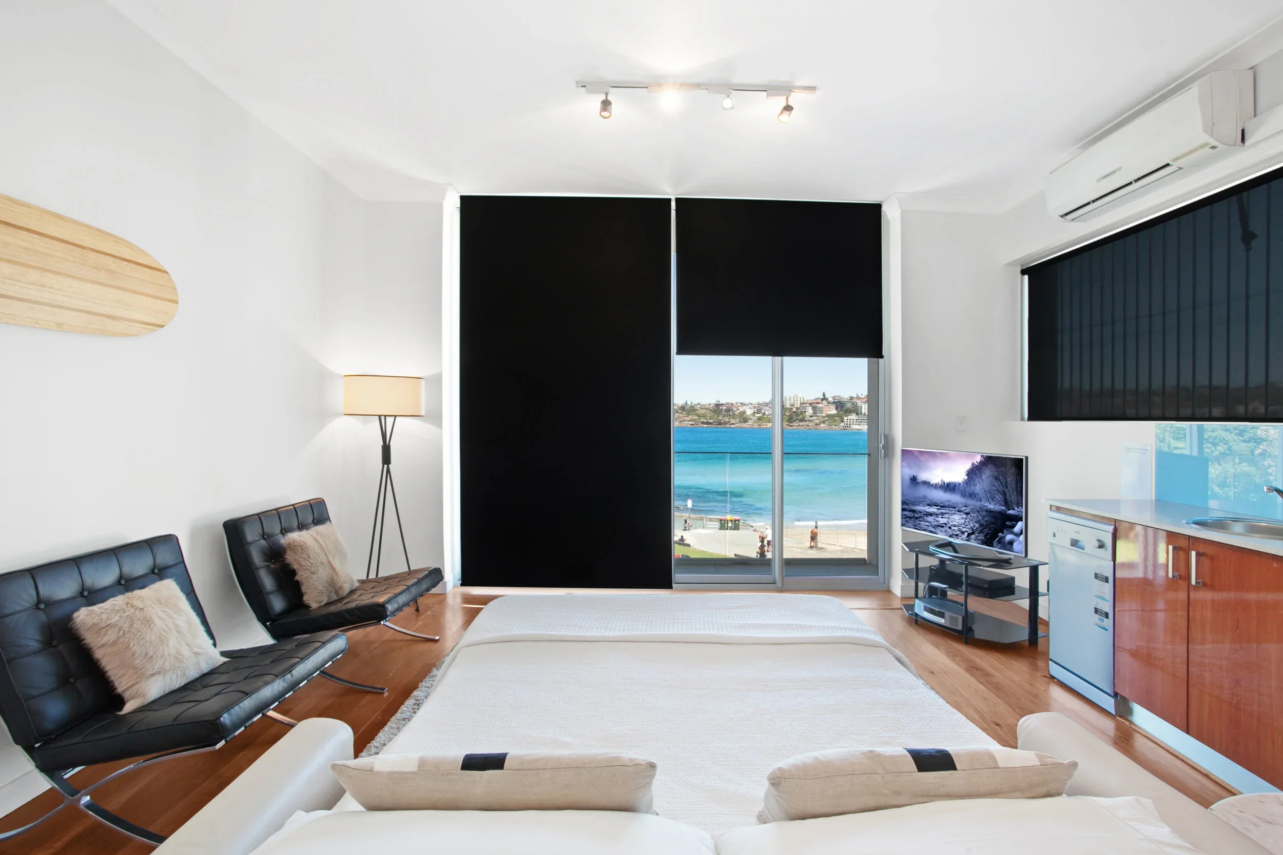 Living room with a large window overlooking a beach, black window coverings, a TV, a small kitchenette, a beige bed, black leather chairs, a standing lamp, and a wooden floor.