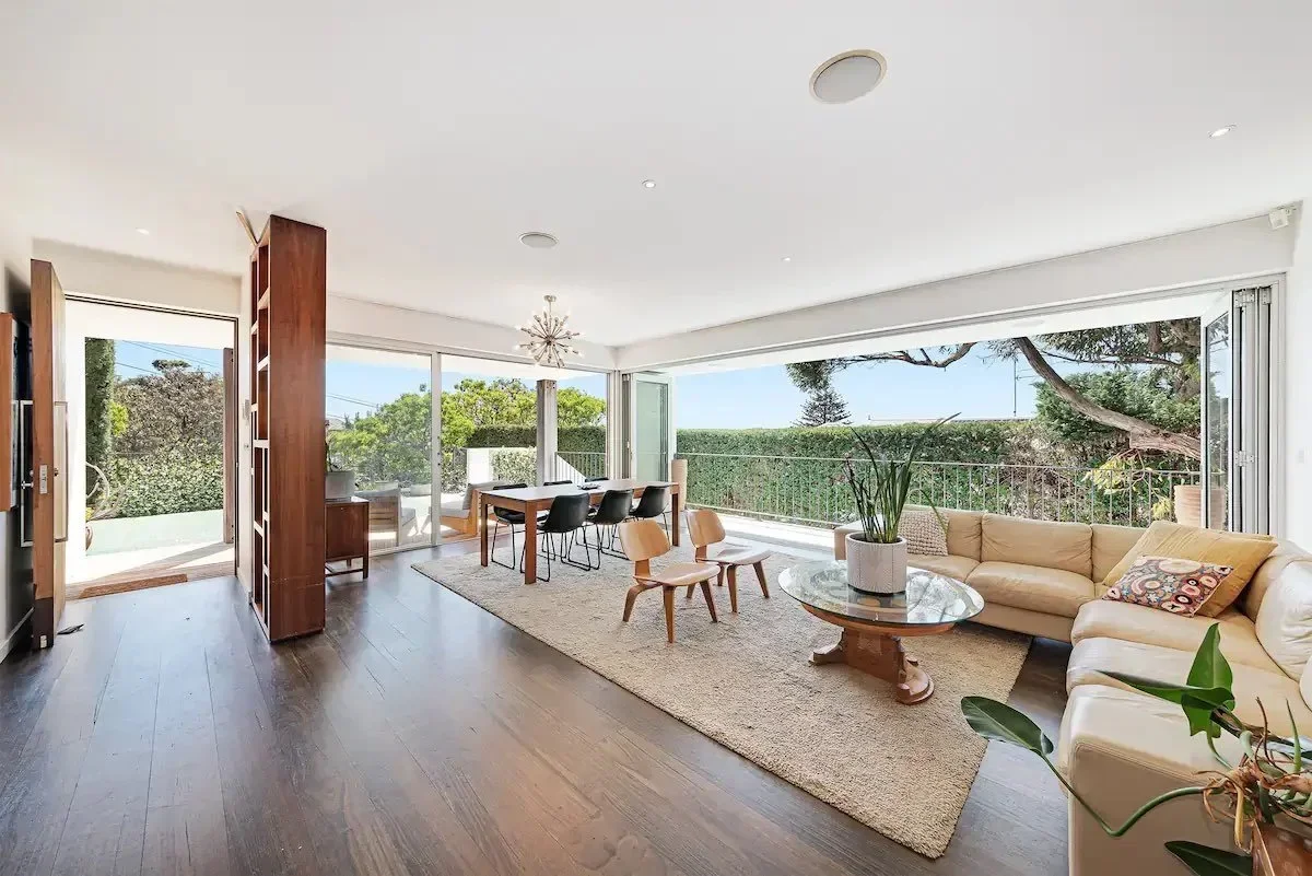 Bright living room with large windows and sliding doors opening to a balcony, featuring a beige sectional sofa, wooden coffee table with a potted plant, dining table with black and wooden chairs, and wooden flooring.