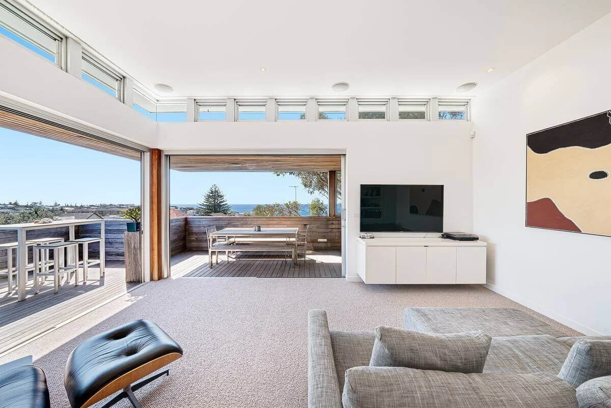 Living room with large sliding glass doors leading to a balcony with outdoor seating, view of trees and ocean, flat-screen TV on white cabinet, abstract wall art, and gray couch.