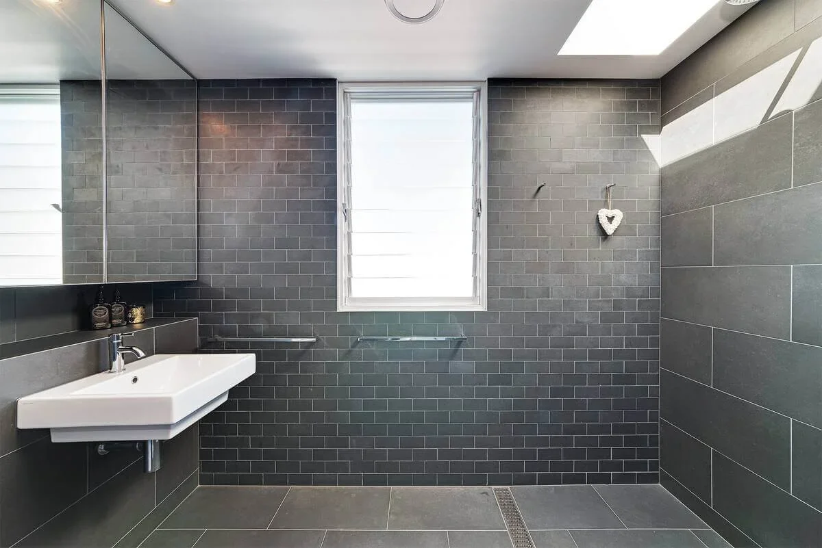 Modern bathroom with black tiled walls, white sink, mirrored cabinet, and window with blinds, minimal decor.