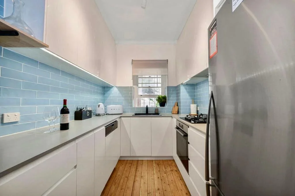 A modern kitchen with white cabinets, a blue tile backsplash, a window above the sink, and various appliances including a toaster, kettle, dishwasher, and stove. There is a bottle of wine and two glasses on the counter.