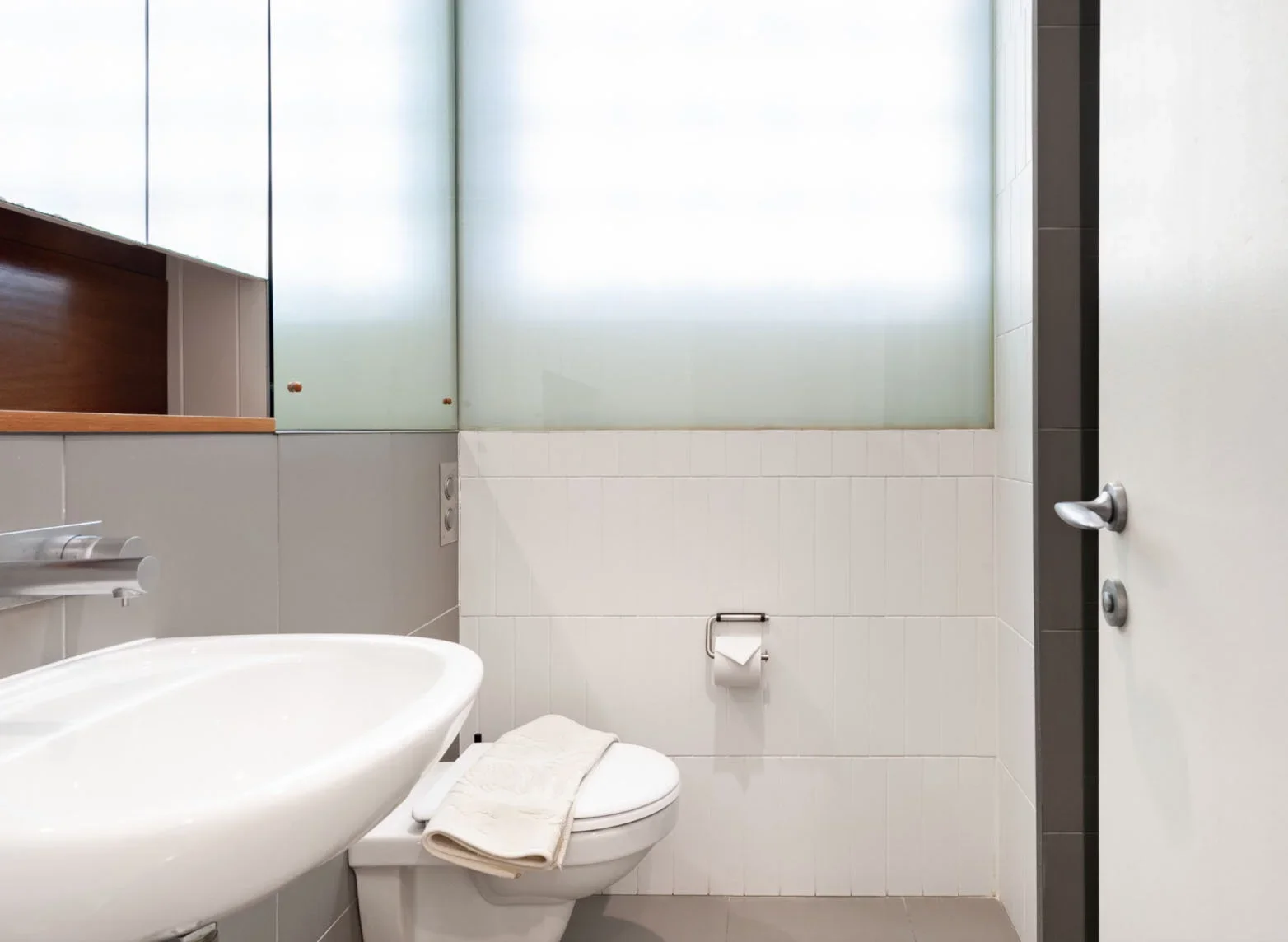 Frosted glass window in a modern bathroom with white tiled walls, a toilet with a folded towel on top, and a white sink. A mirror is partially visible on the left, and a door with an open door handle is on the right.