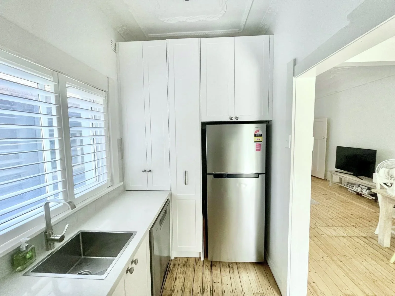 View of a kitchen with white cabinets, a stainless steel refrigerator, a white countertop with a sink, a window with blinds, and a glimpse into a living room with a TV and wooden floor.