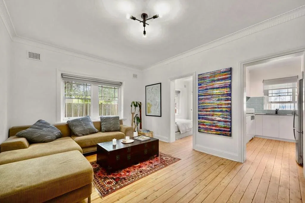 Living room with beige sectional sofa, gray pillows, wooden coffee table, patterned rug, white walls, art on the wall, window with blinds, doorway to a bedroom with white bedding, doorway to the kitchen with white cabinets and gray backsplash, hardwo