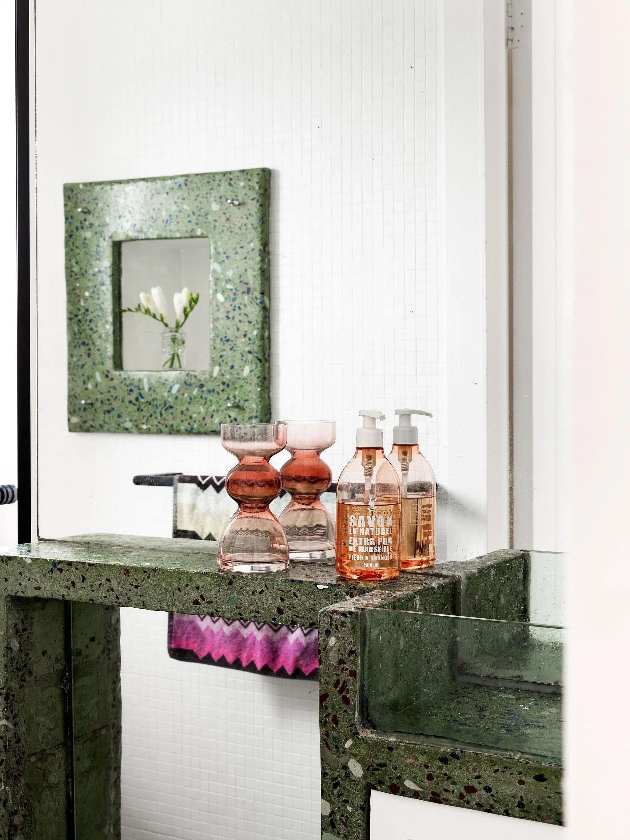 A bathroom vanity with a green terrazzo surface holding a pair of pink glass candle holders and two pink soap dispensers, a mirror with a green terrazzo frame, and a pink and black striped towel hanging beneath the surface.