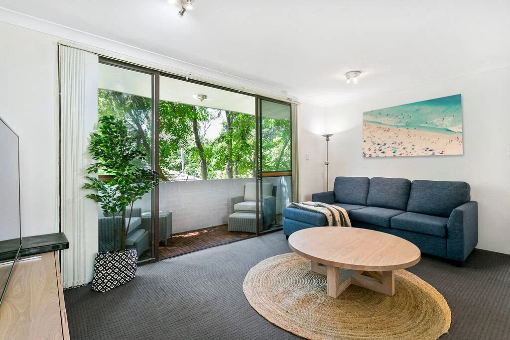 Living room with a large blue sofa, a wooden coffee table on a circular rug, a tall floor lamp, a painting of a beach scene, a sliding glass door leading to a balcony with outdoor chairs, and a potted plant near the sliding door.