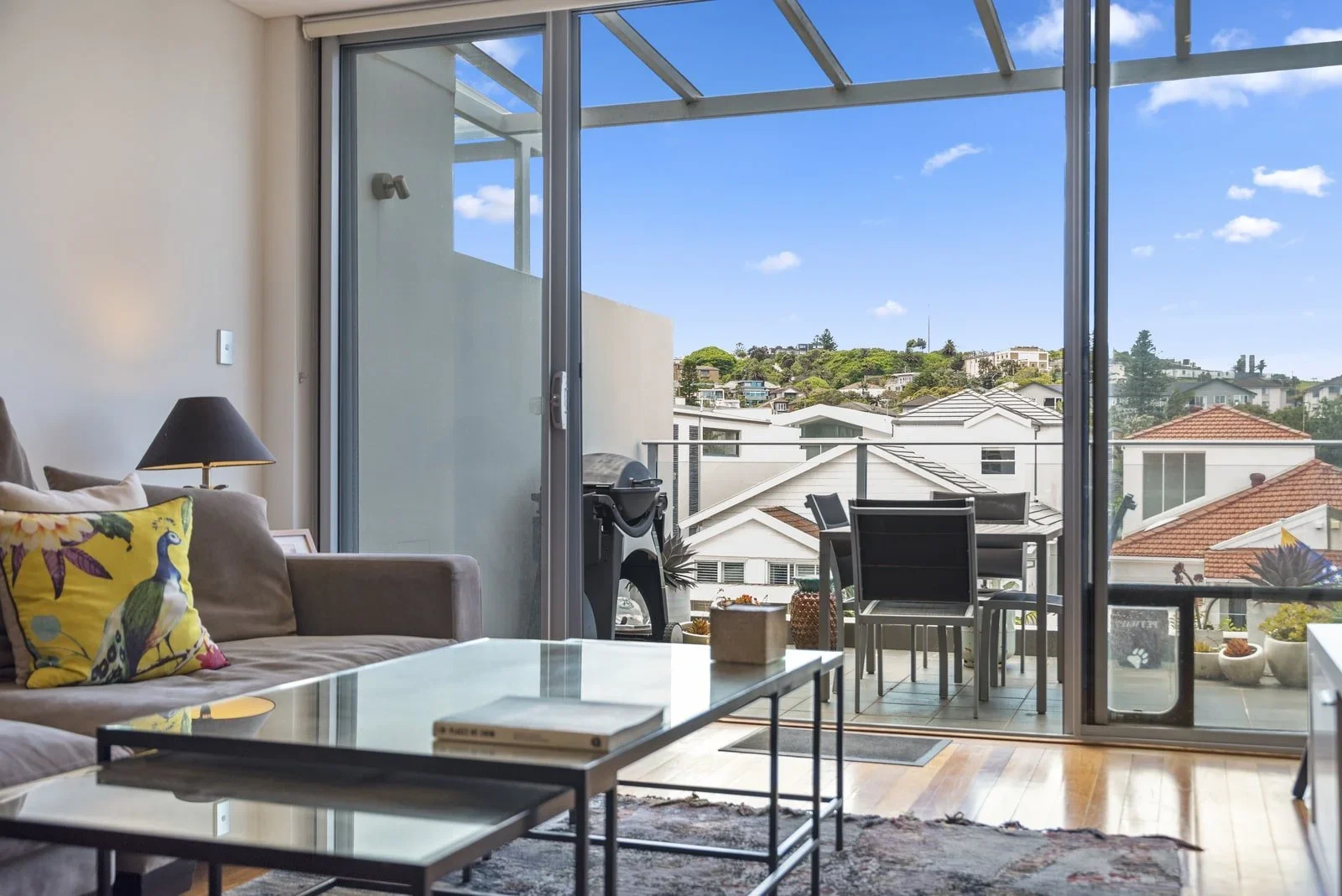 Living room with glass sliding door leading to balcony, outdoor table with chairs, barbecue grill, cityscape with houses and hills in the background, and a colorful pillow on a beige sofa.