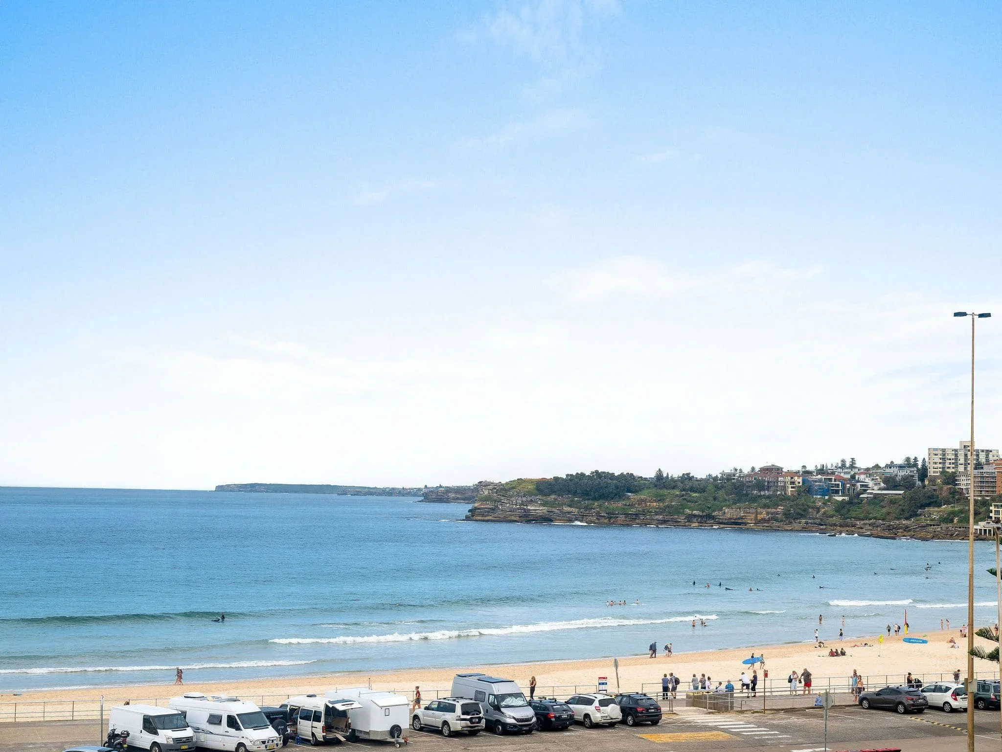 Beach with people, cars, and vans parked along the boardwalk. Ocean waves, rocky cliffs, and buildings in the background under a partly cloudy sky.