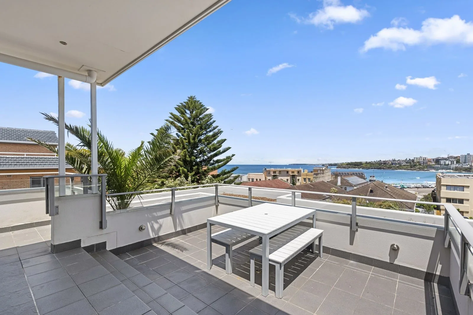 Ocean view balcony with white outdoor furniture, trees, and buildings under a blue sky with clouds.