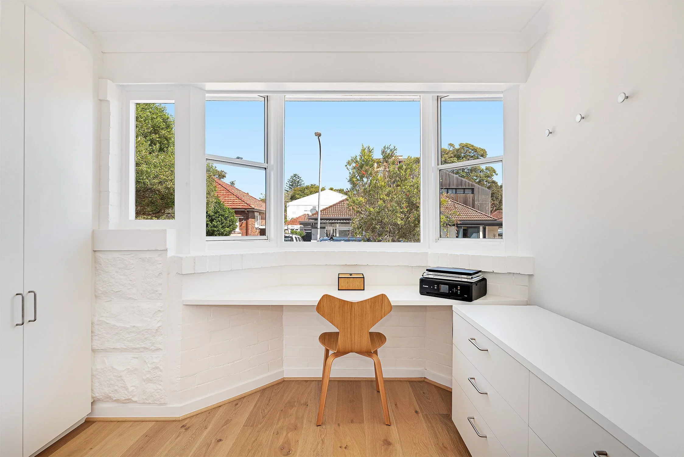 A minimalist home office corner with a wooden chair, a built-in white desk under a large window, and a black printer on the right side of the desk in a bright room with hardwood floors.