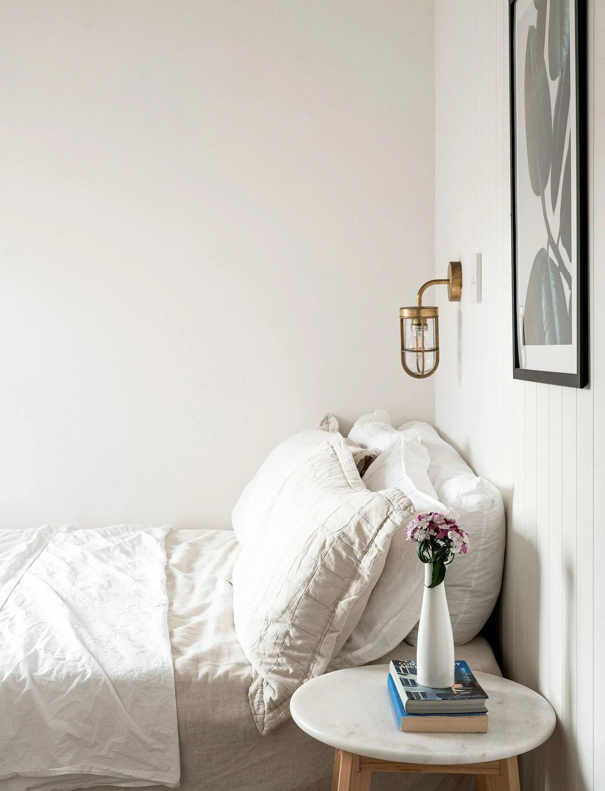 A cozy bedroom with a white bed, beige pillows, a small round side table with a vase of pink flowers, a book, and a framed abstract wall art.