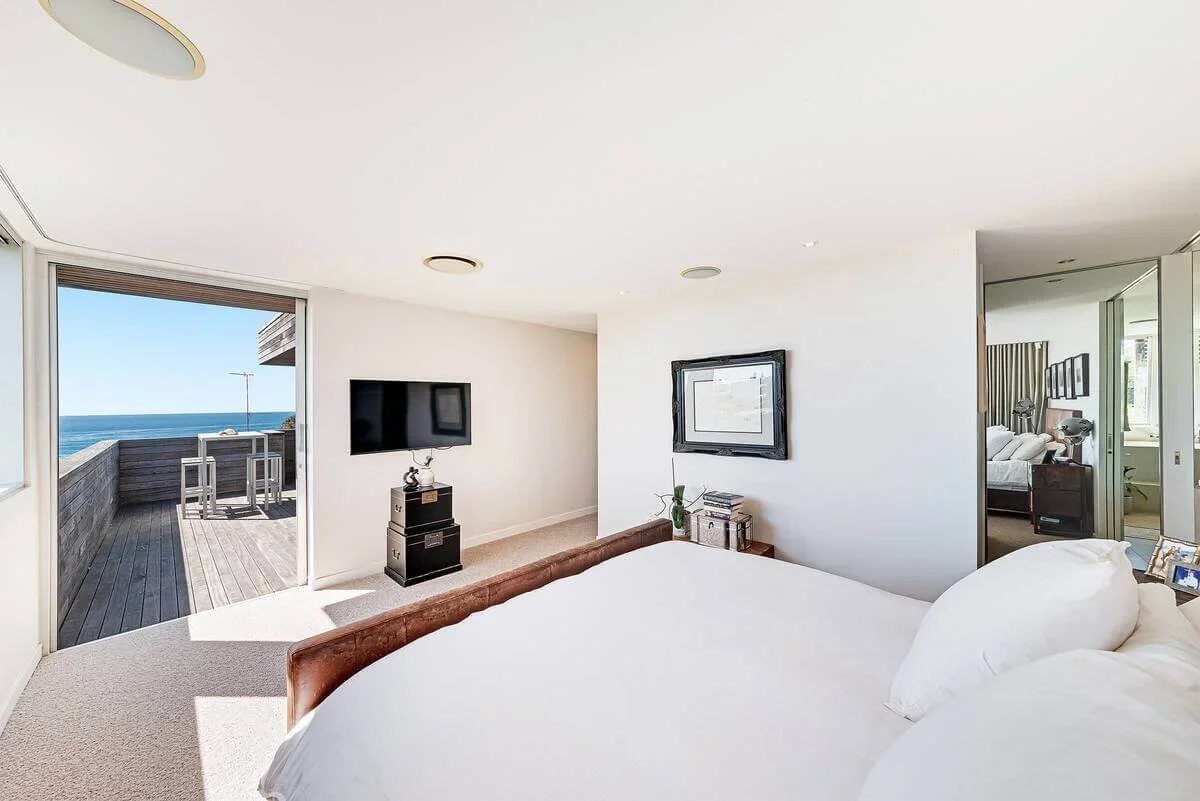 Bright bedroom with a large bed, balcony view of ocean, and a small outdoor seating area.