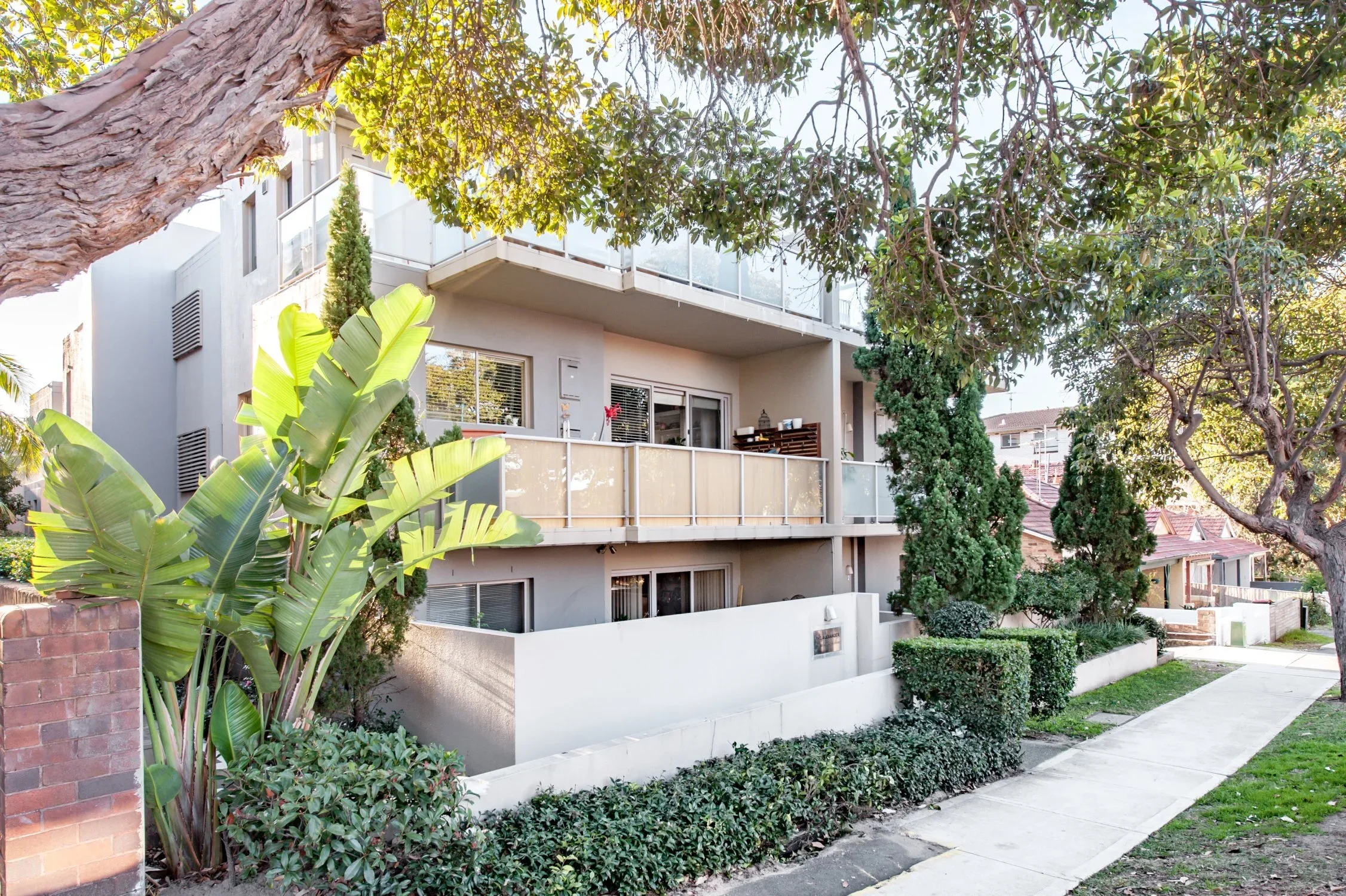 Apartment building with balconies, surrounded by green trees and shrubs, along a sidewalk in a residential neighborhood.