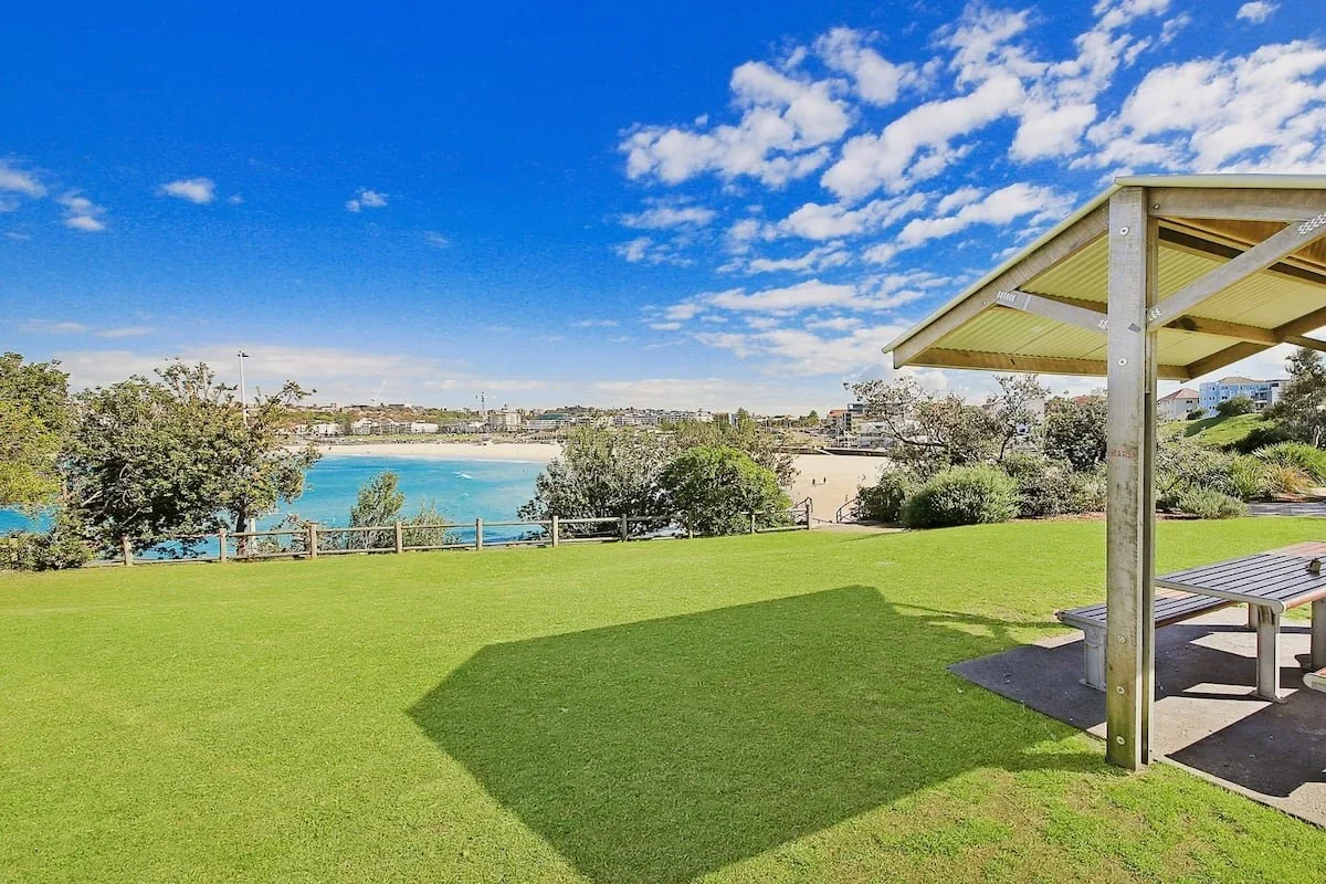 Sunlit park with green grass, a shaded picnic table, trees, and a view of a blue river or bay with a sandy beach, under a partly cloudy sky.