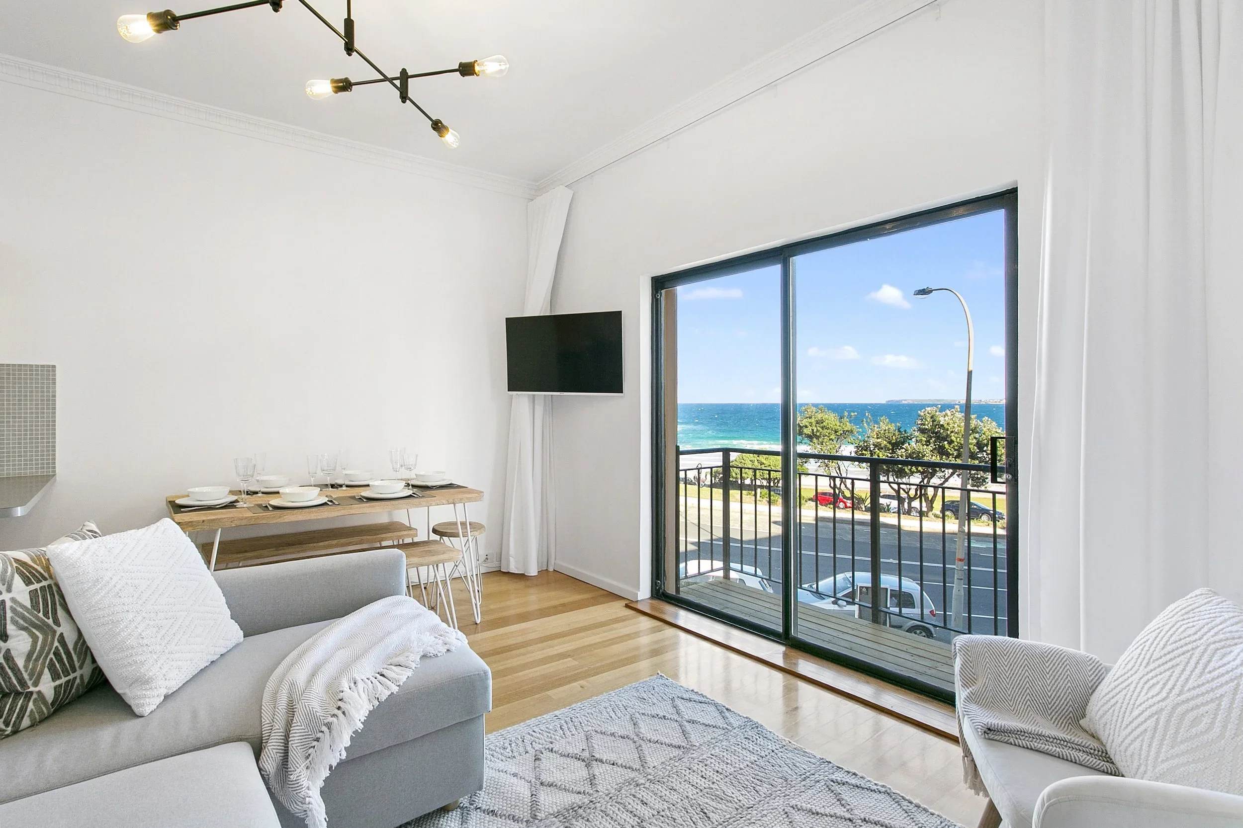 Living room with a large sliding glass door overlooking the ocean, white walls, wooden flooring, a corner sofa with throw pillows and blanket, a wooden dining table with chairs, a flat-screen TV, and a ceiling light fixture.