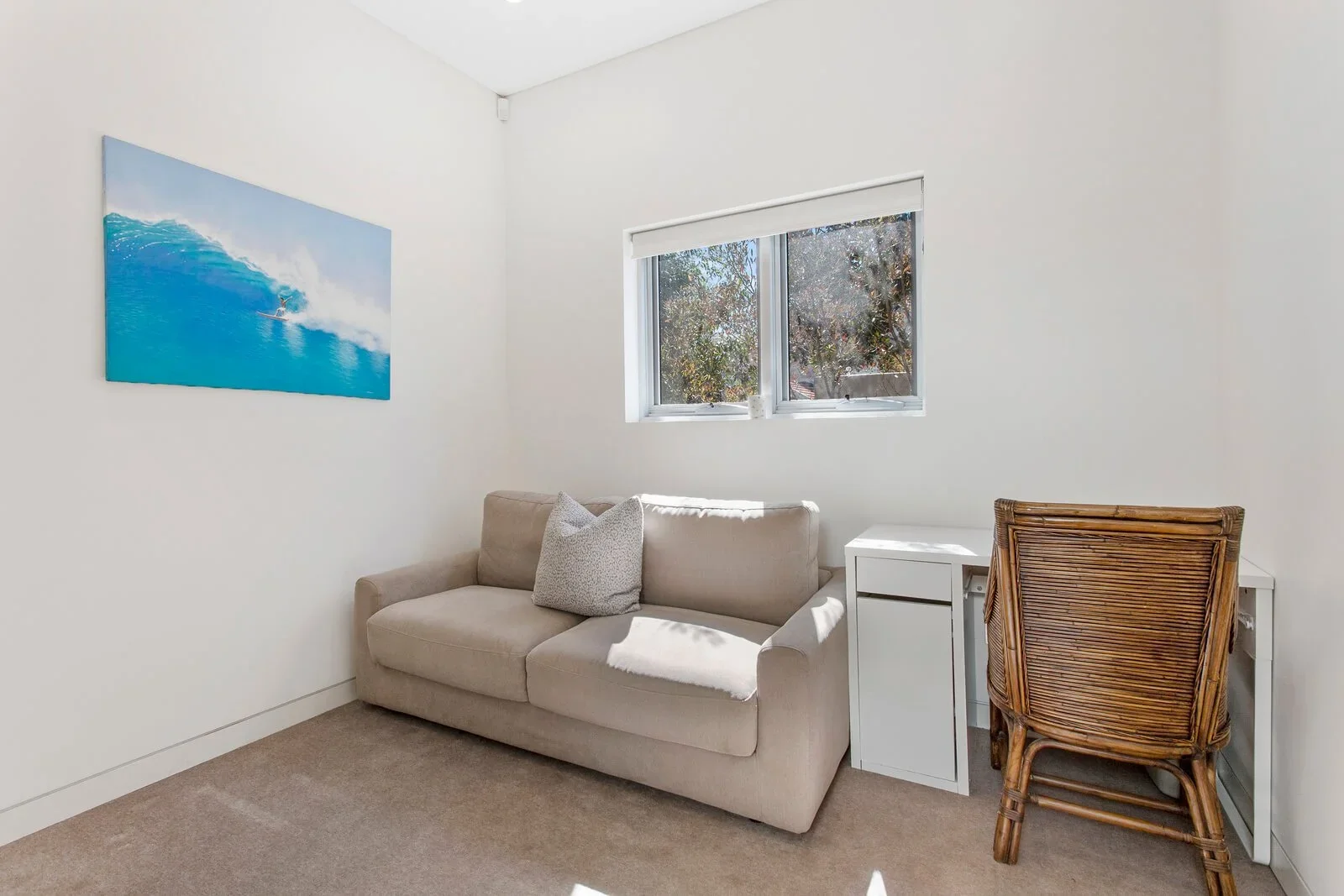 A beige sofa with a patterned pillow, a small white side table, and a wooden chair in a room with white walls. There is a window with trees outside and a wall art of a surfer riding a wave.