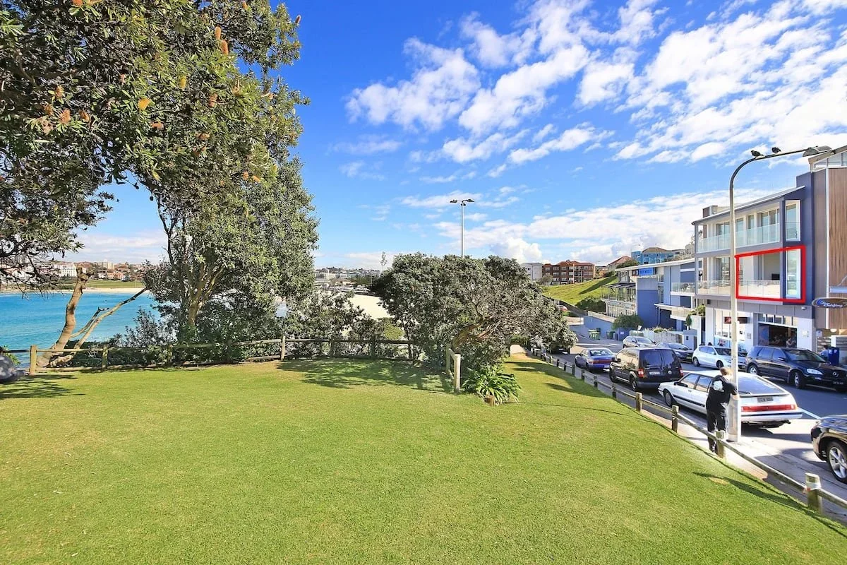 A grassy park area with trees, overlooking a body of water. Adjacent to the park is a street with parked cars and modern residential buildings, under a partly cloudy sky.