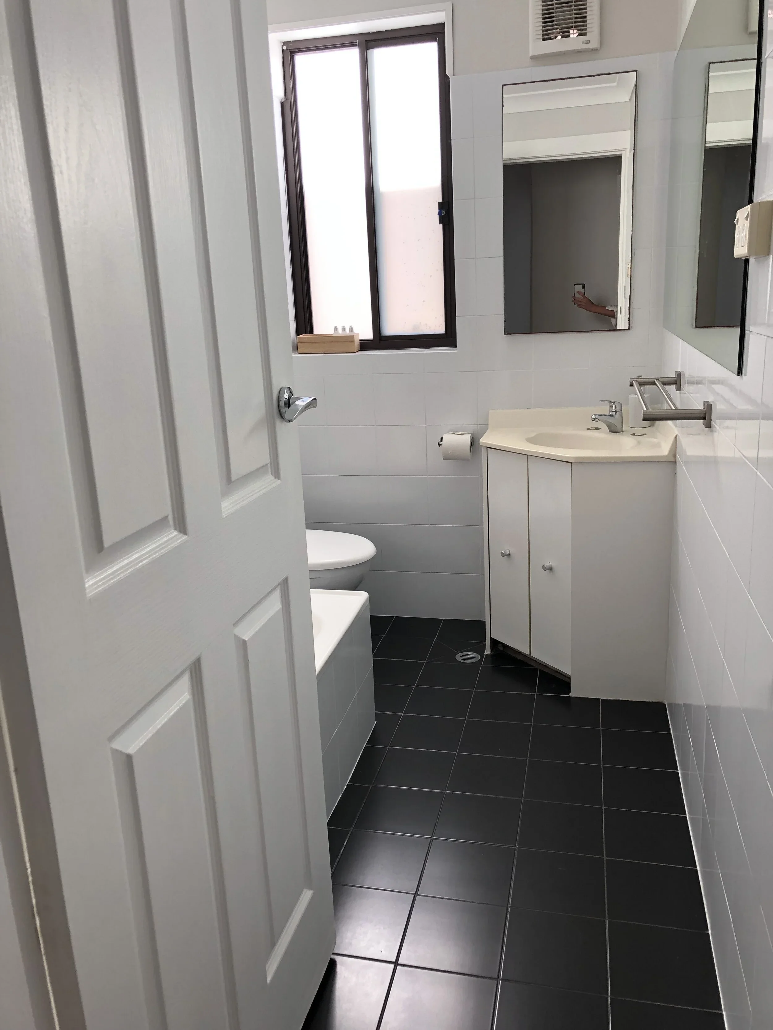 Small bathroom with black tile flooring, white walls, a small white vanity with a sink, a mirror, a window, a toilet, a bathtub, and a partially open door.