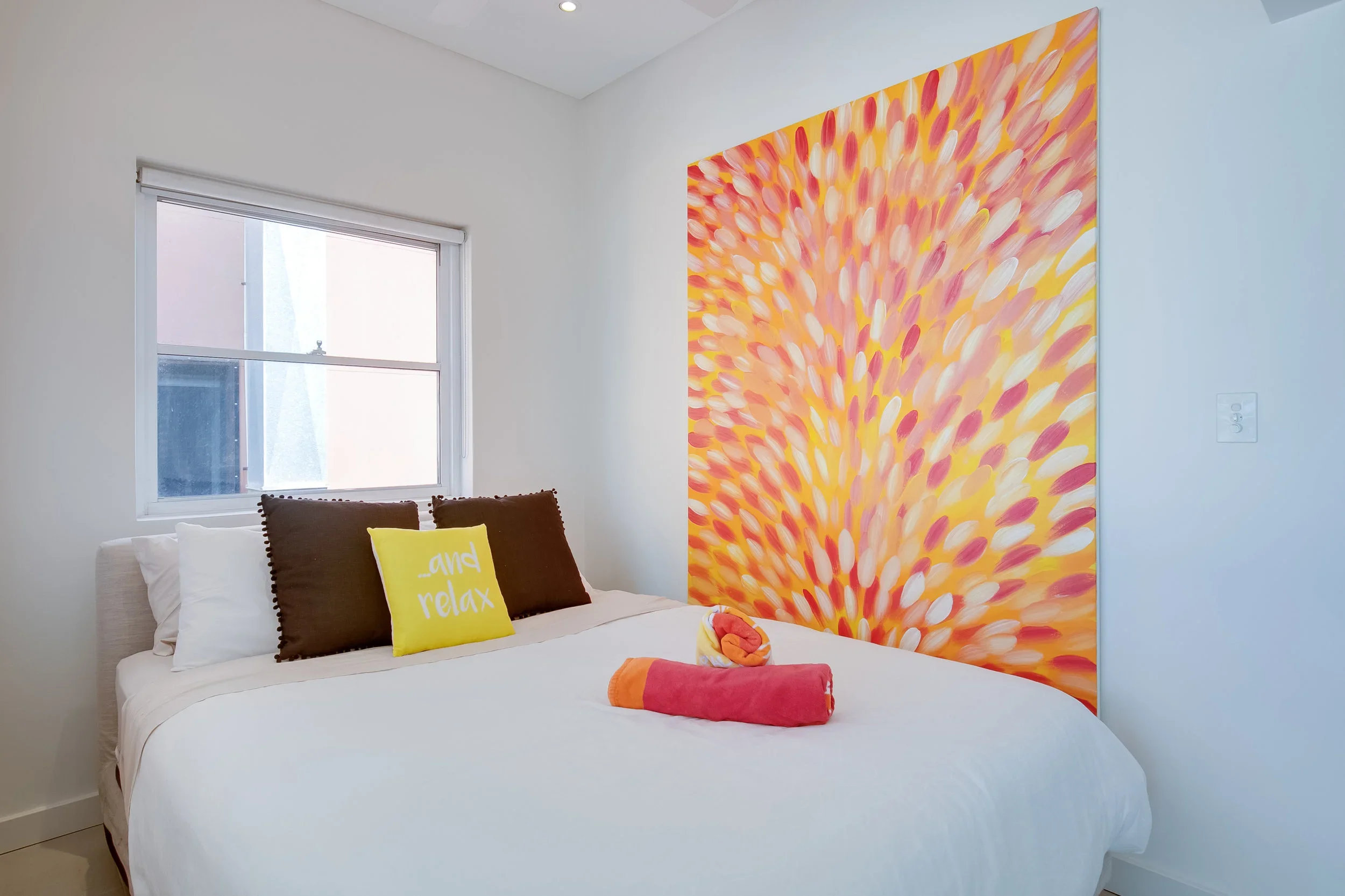 A bedroom with white walls and a large abstract painting with warm colors on the wall behind the bed. The bed has white linens, two dark brown pillows, a yellow pillow with white text that reads 'and relax,' and a rolled orange towel on top.