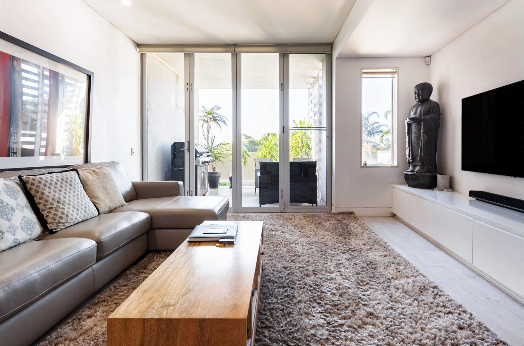 Modern living room with beige leather sectional sofa, wooden coffee table, shaggy beige rug, and a flat-screen TV mounted on white wall. Large glass sliding doors lead to a balcony with outdoor seating and palm trees.
