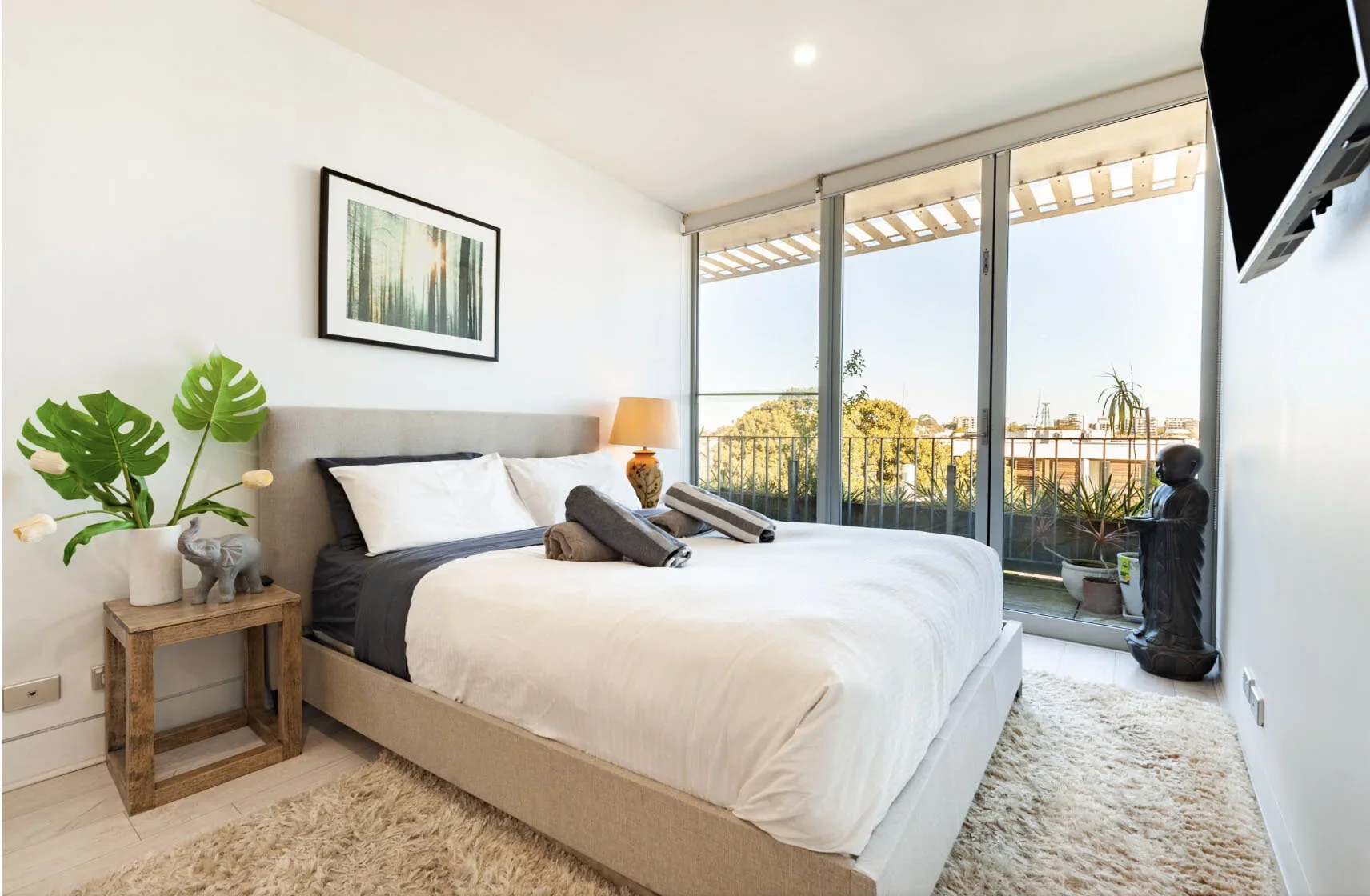Modern bedroom with large windows, a bed with white and black bedding, a wooden nightstand with a plant and elephant sculpture, a table lamp, a wall-mounted TV, and decorative statues, with a view of trees and buildings outside.